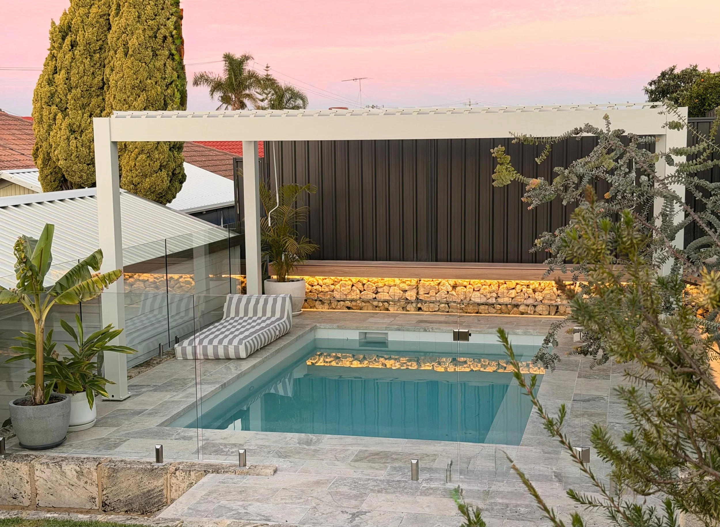Residential backyard with pool, surrounding plants, a lounge chair with striped cushions, and a pergola at sunset with pink sky.