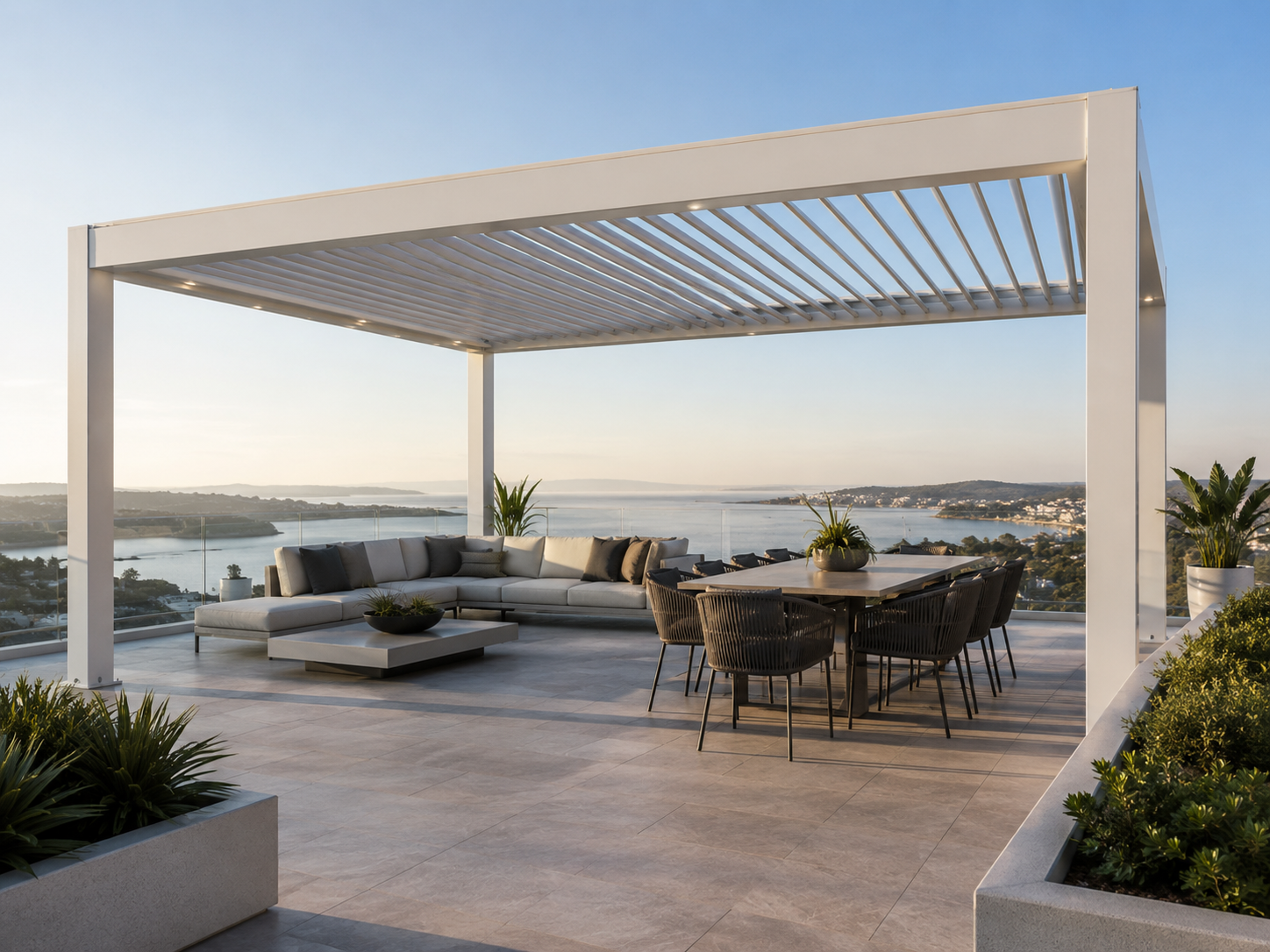 Luxury outdoor terrace with modern white pergola, sectional sofa with cushions, dining table with chairs, potted plants, overlooking a body of water and landscape at sunset.