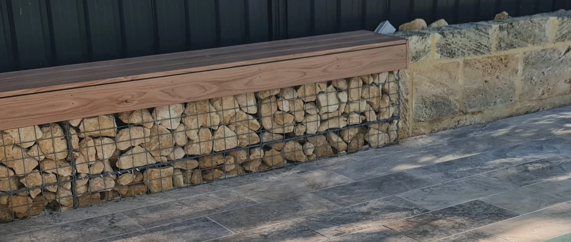 Image showing a decorative stone gabion wall , topped with a oak composite decking benchseat, against a dark metal fence and silver travertine paving