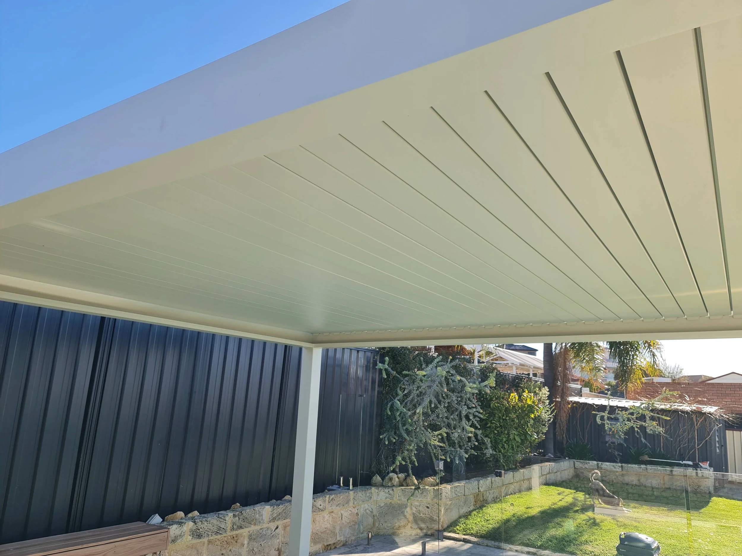 Backyard with a bioclimatic pergola roof, dark metal fence, garden bed with plants, and a small dog standing on the grass.