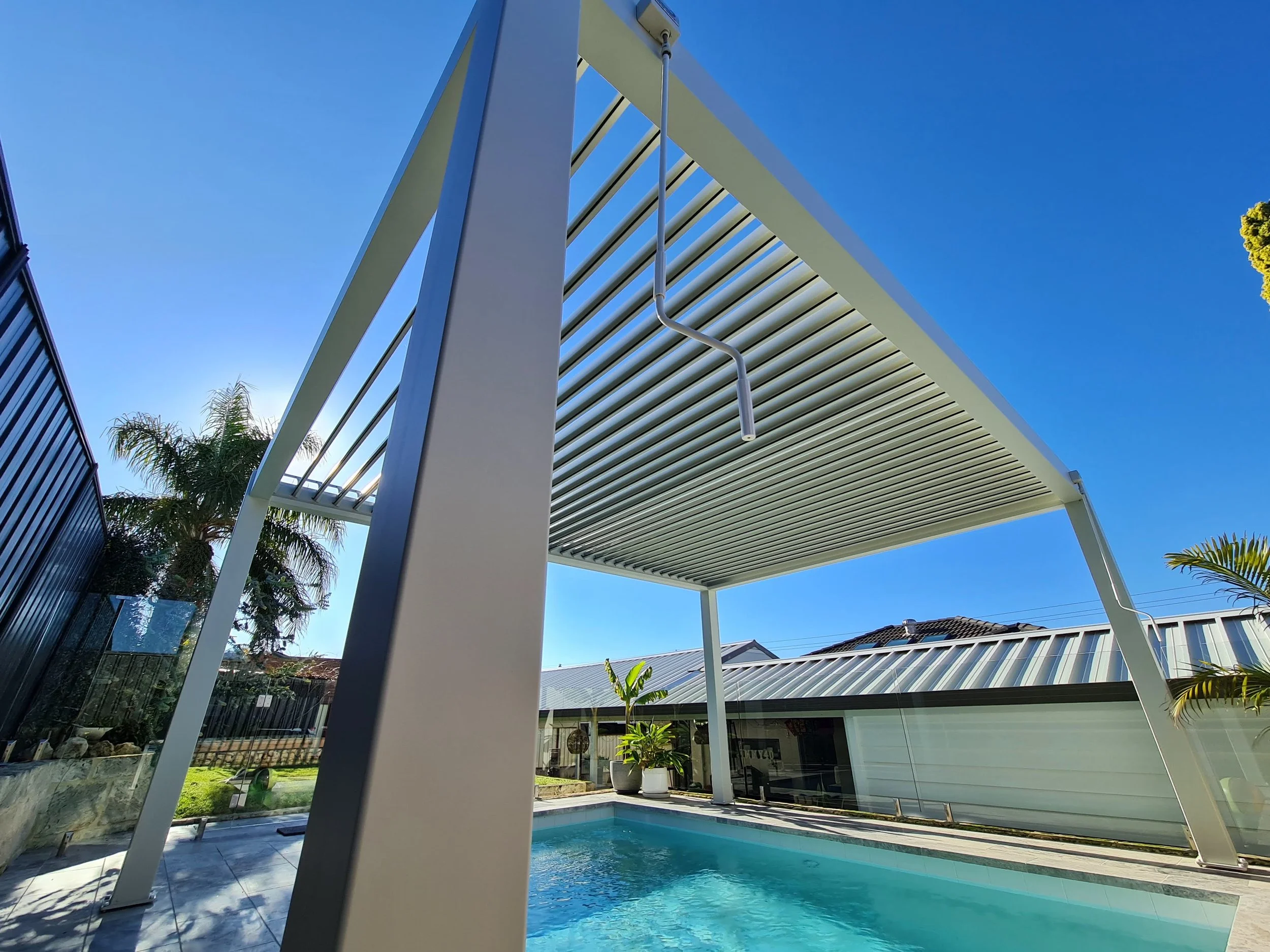 Modern backyard with a pool, covered seating area with white slatted roof, and green trees; clear blue sky.