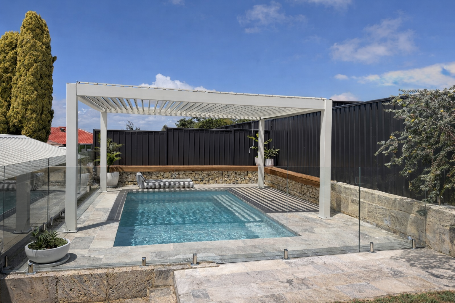 Backyard with a small rectangular swimming pool, surrounded by stone tiles, with barley grass and fence around it. There is a white pergola over the pool, with striped outdoor furniture and potted plants nearby. The sky is partly cloudy with blue.