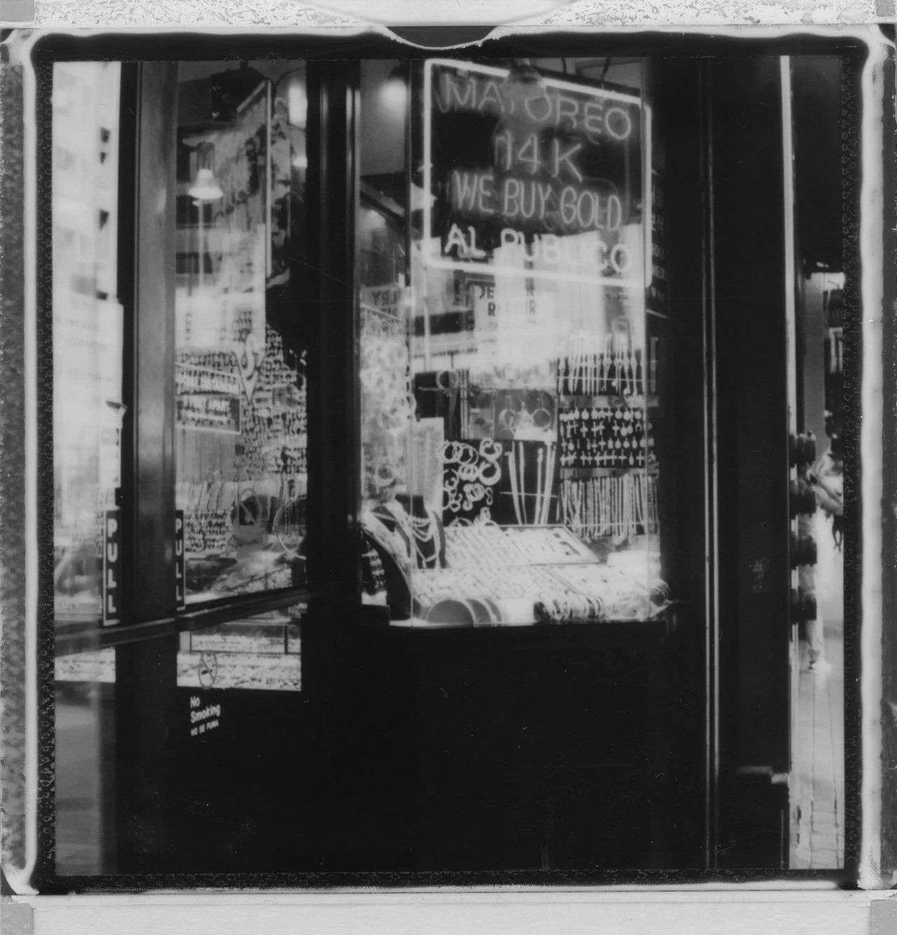 Storefront display with neon signs advertising 'MAYOREO 14K', 'WE BUY GOld', and other text, with various items on display inside, including jewelry and decorative objects, and a reflection of street scenes outside.