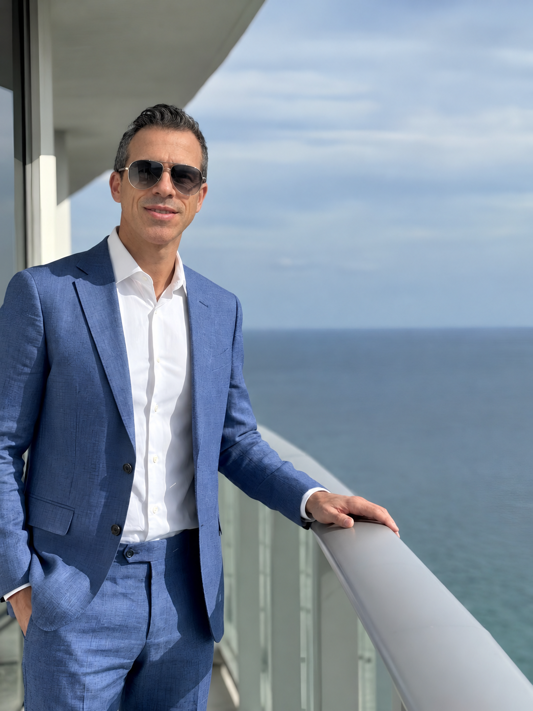 A man in a blue suit and white shirt standing on a balcony overlooking the ocean, wearing sunglasses.