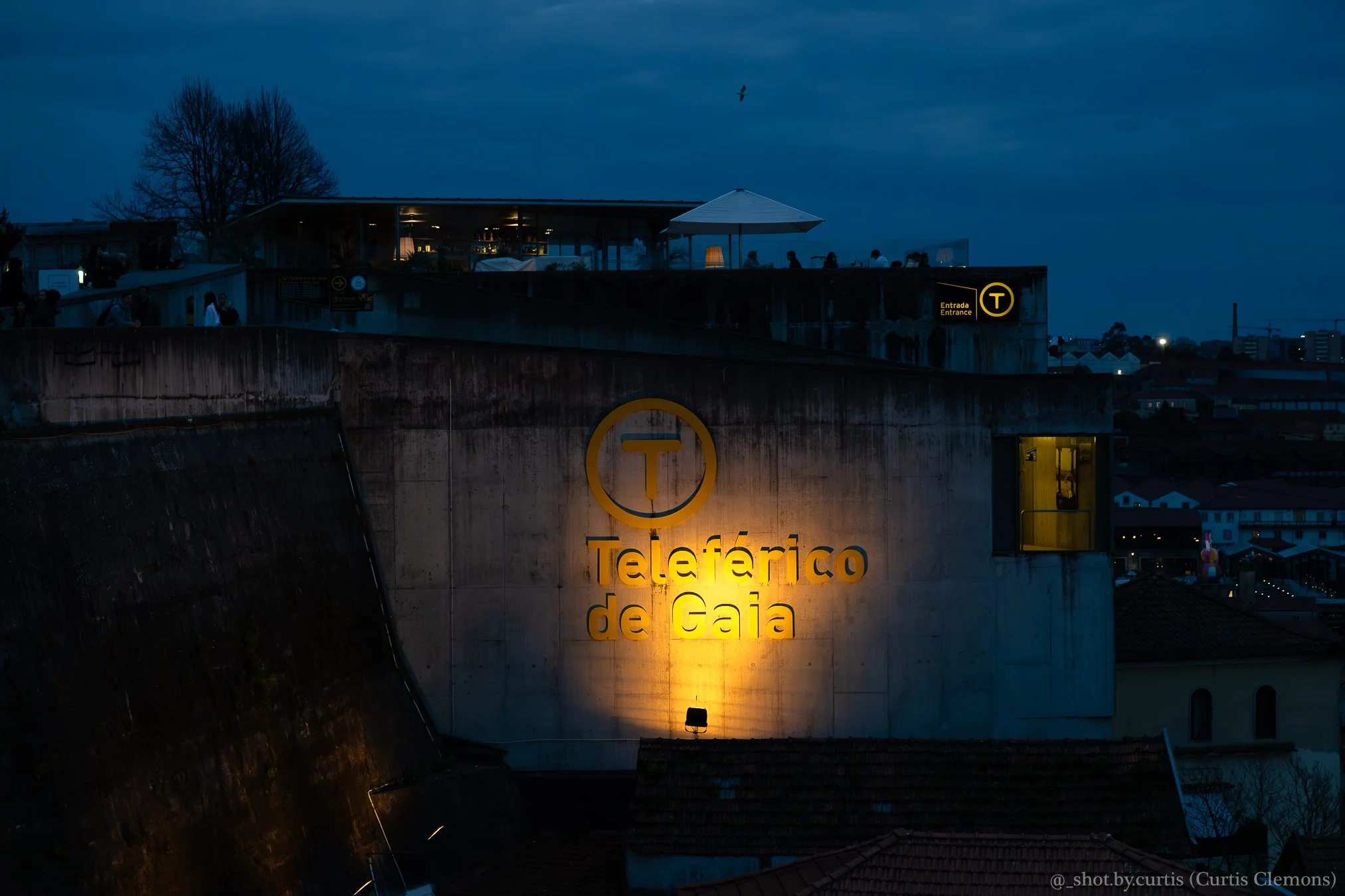 Nighttime city view showing the Teleférico de Gaia cable car station with illuminated signs and people inside, against a dark sky with a few clouds and a bird flying overhead.