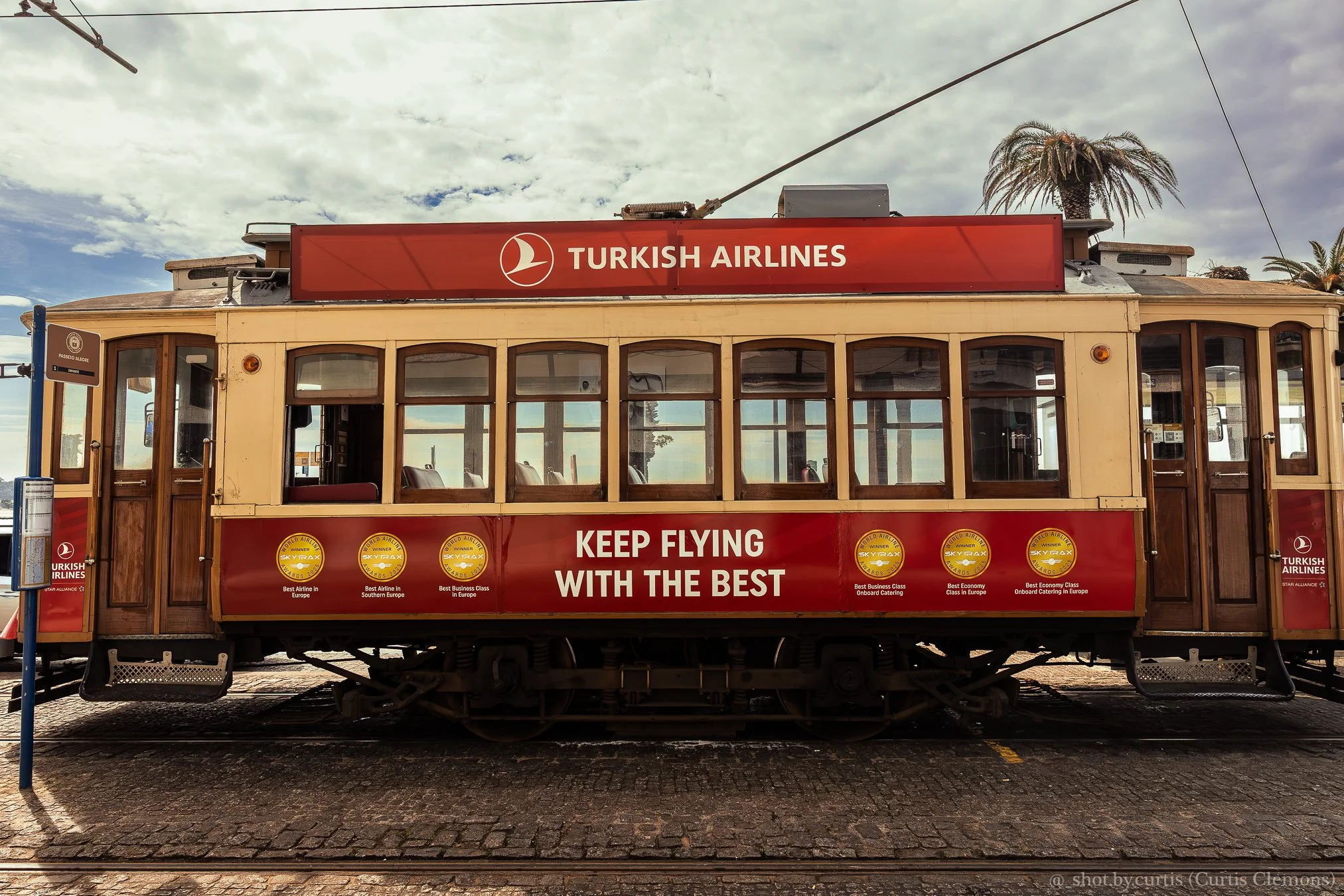 A vintage tram with a red sign that reads 'Turkish Airlines' and 'Keep Flying With The Best' on its side. The tram has wooden window frames, a beige body, and is parked on a cobblestone street. There are palm trees and a cloudy sky in the background.