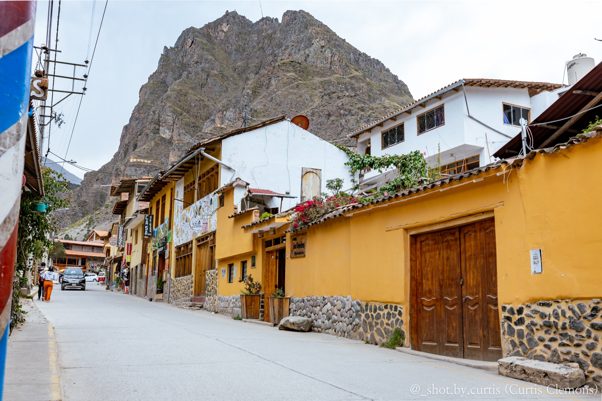 A street scene in a town at the base of a tall rocky mountain, with colorful buildings, some with plants on balconies, and a few people walking.