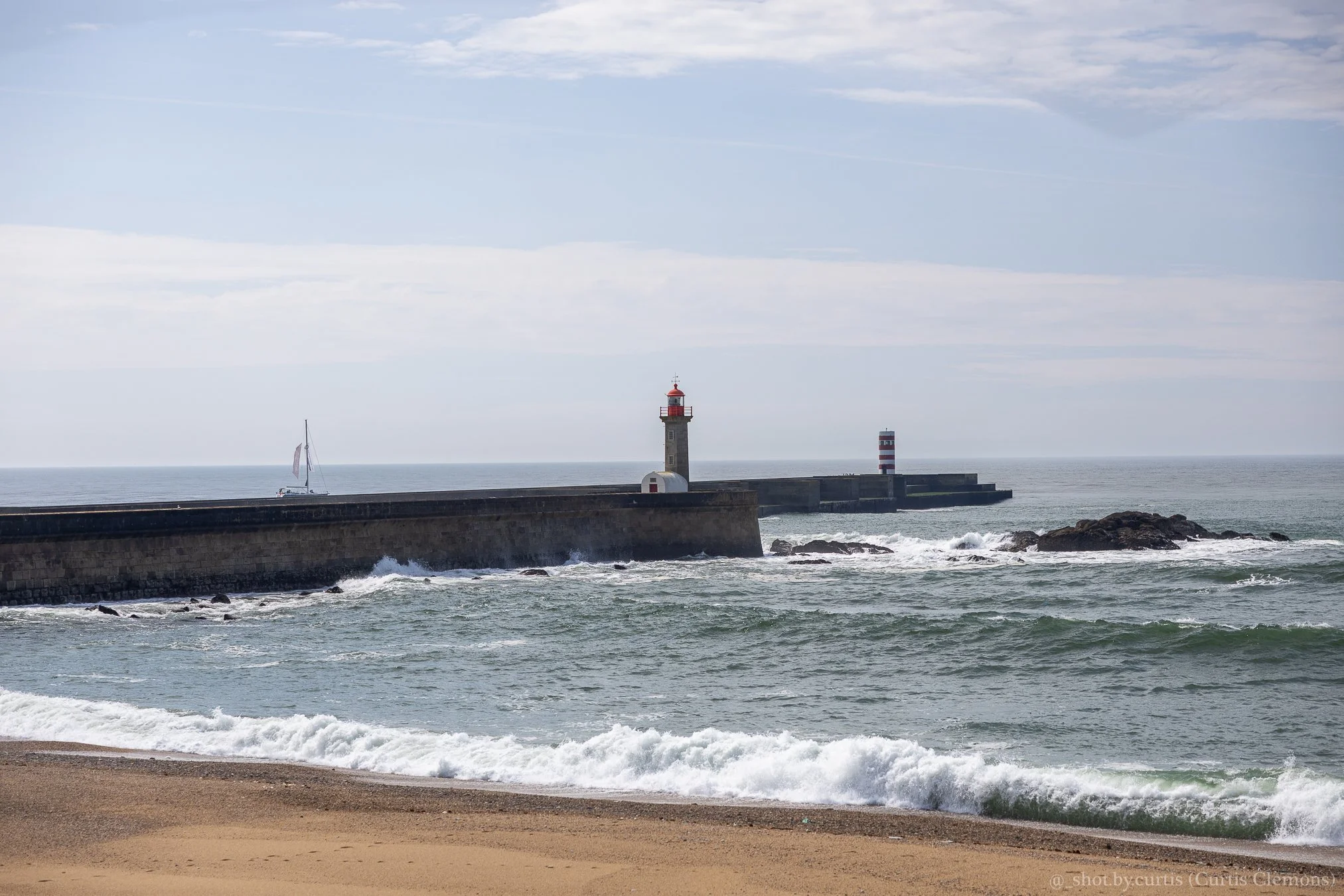 A lighthouse on a breakwater by the ocean with a small sailboat nearby and rocky shoreline, under a cloudy sky.