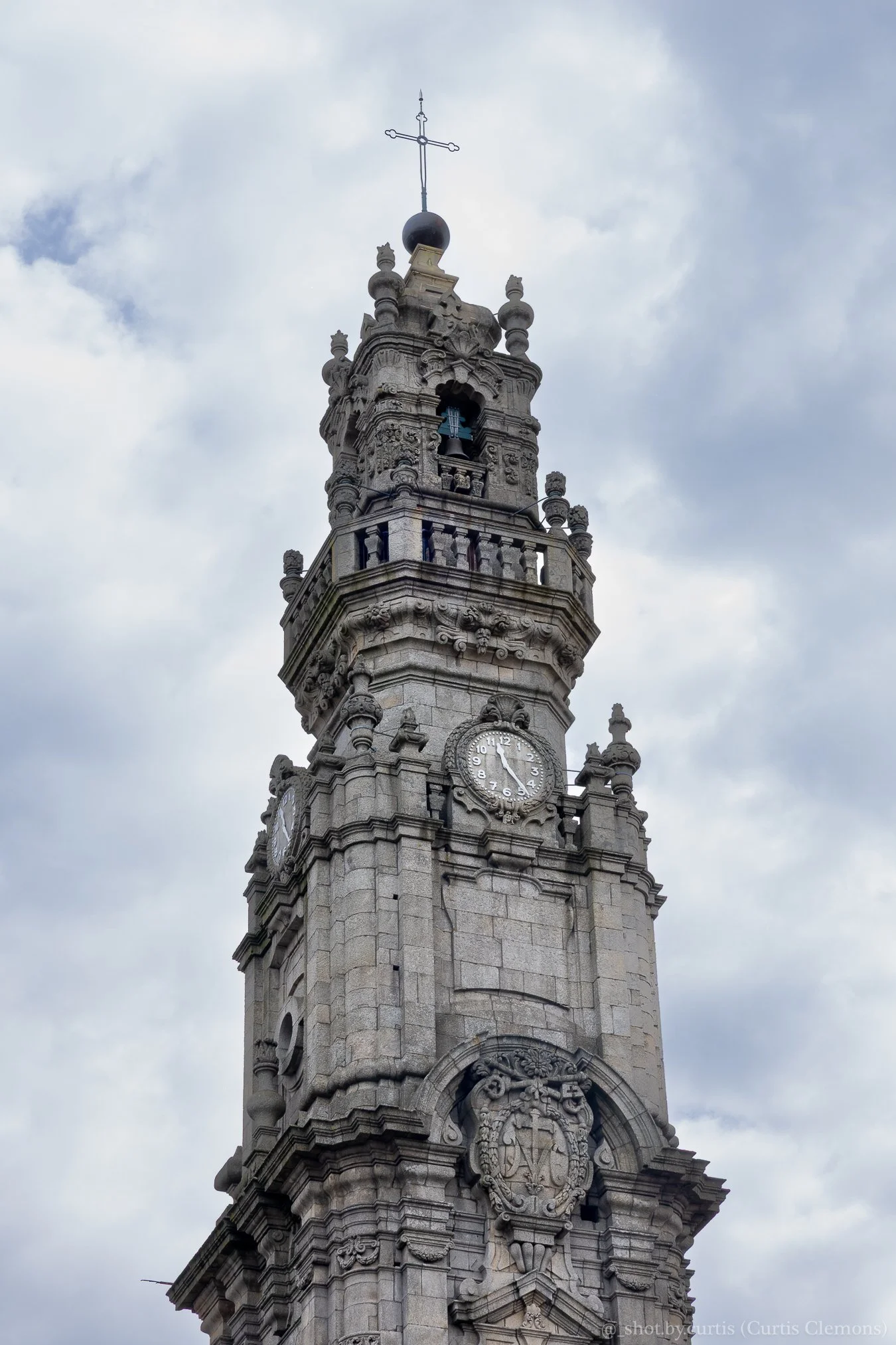 A tall, ornate stone clock tower with intricate carvings, a clock face showing 4:09, and a small bell visible in an open section near the top, topped with a cross against a cloudy sky.