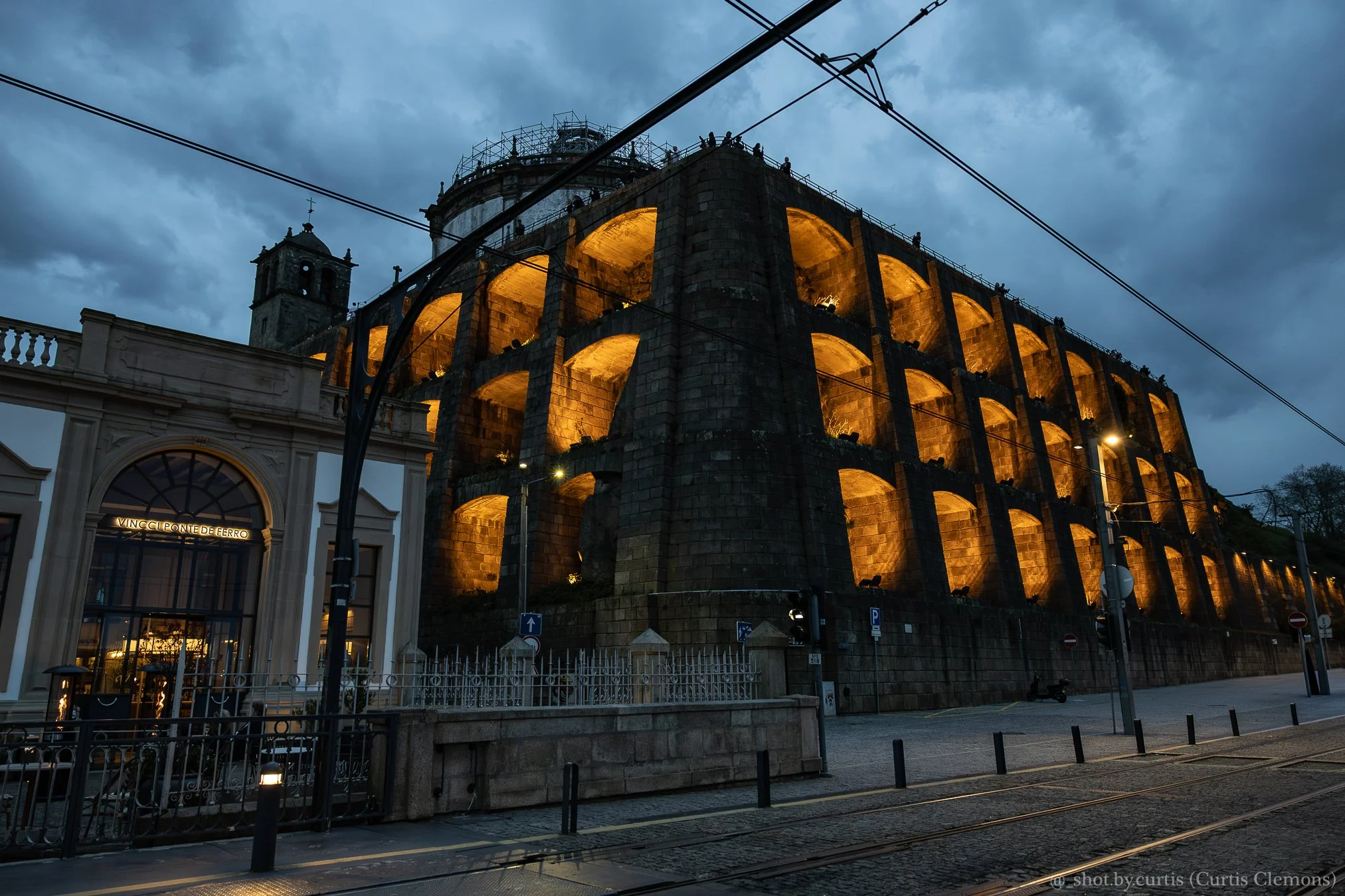 The illuminated ancient Roman coliseum at dusk with a dark, cloudy sky in the background, above a modern city street with tram wires, cars, and an entrance building.
