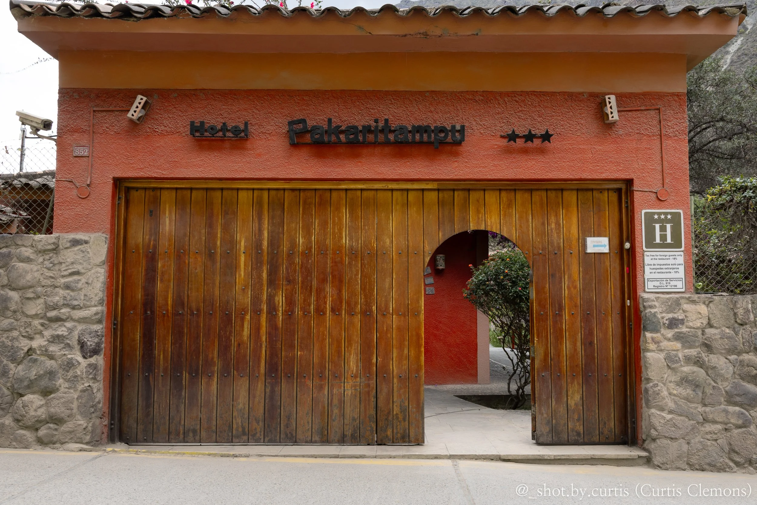 Red stucco building with the words 'Hotel Pakaritampu' and three stars displayed on it, featuring a large wooden gate and stone wall, with surrounding trees and mountains in the background.