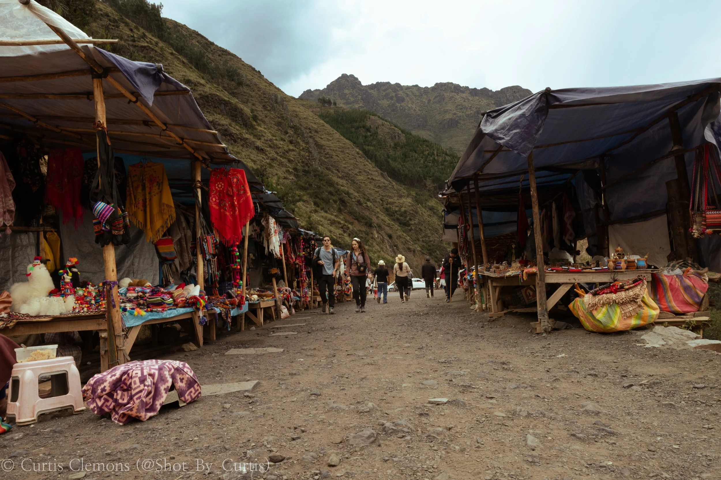 Outdoor market with stalls selling colorful textiles and handmade crafts, set against a mountainous landscape and cloudy sky. Several people browse and walk between the stalls on a dirt ground.