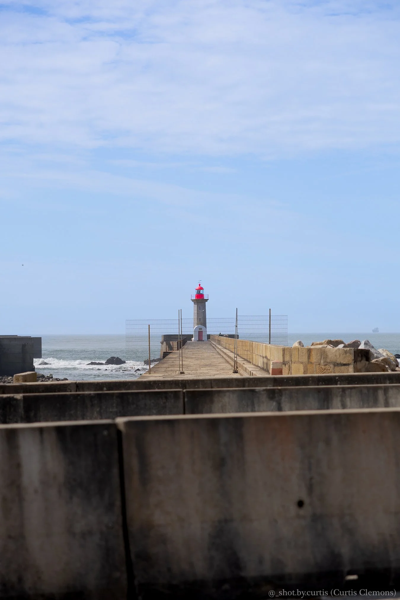 A lighthouse at the end of a breakwater with a cloudy sky above and the ocean in the background, viewed through a weathered wooden barrier.