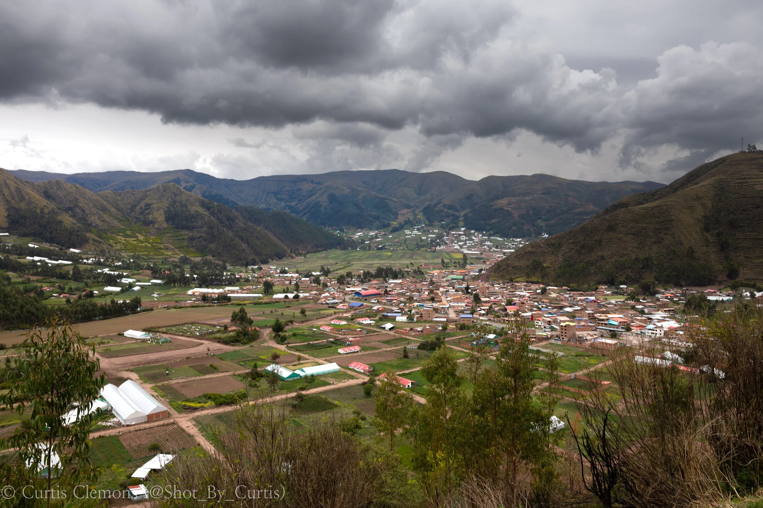 A wide view of a mountainous landscape with a small town or village nestled in a valley. The mountains are green and brown, under a cloudy, dark sky with heavy gray clouds.