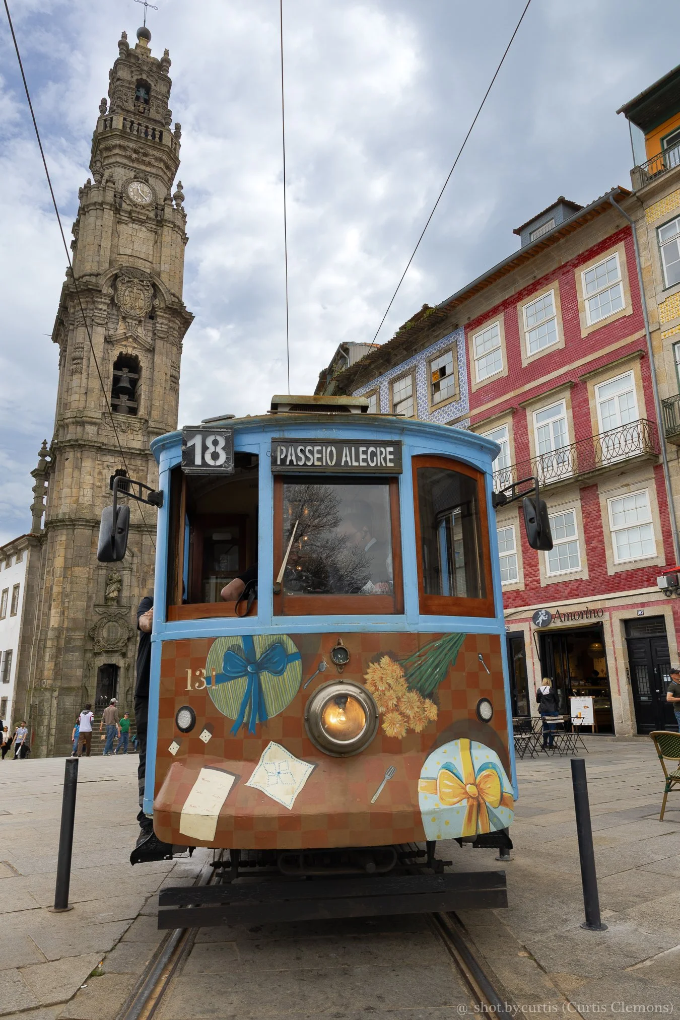 A classic blue tram with colorful decorations, including gift-wrapped presents and bows, traveling on tracks in a city square. In the background, there is a historical clock tower and colorful buildings.