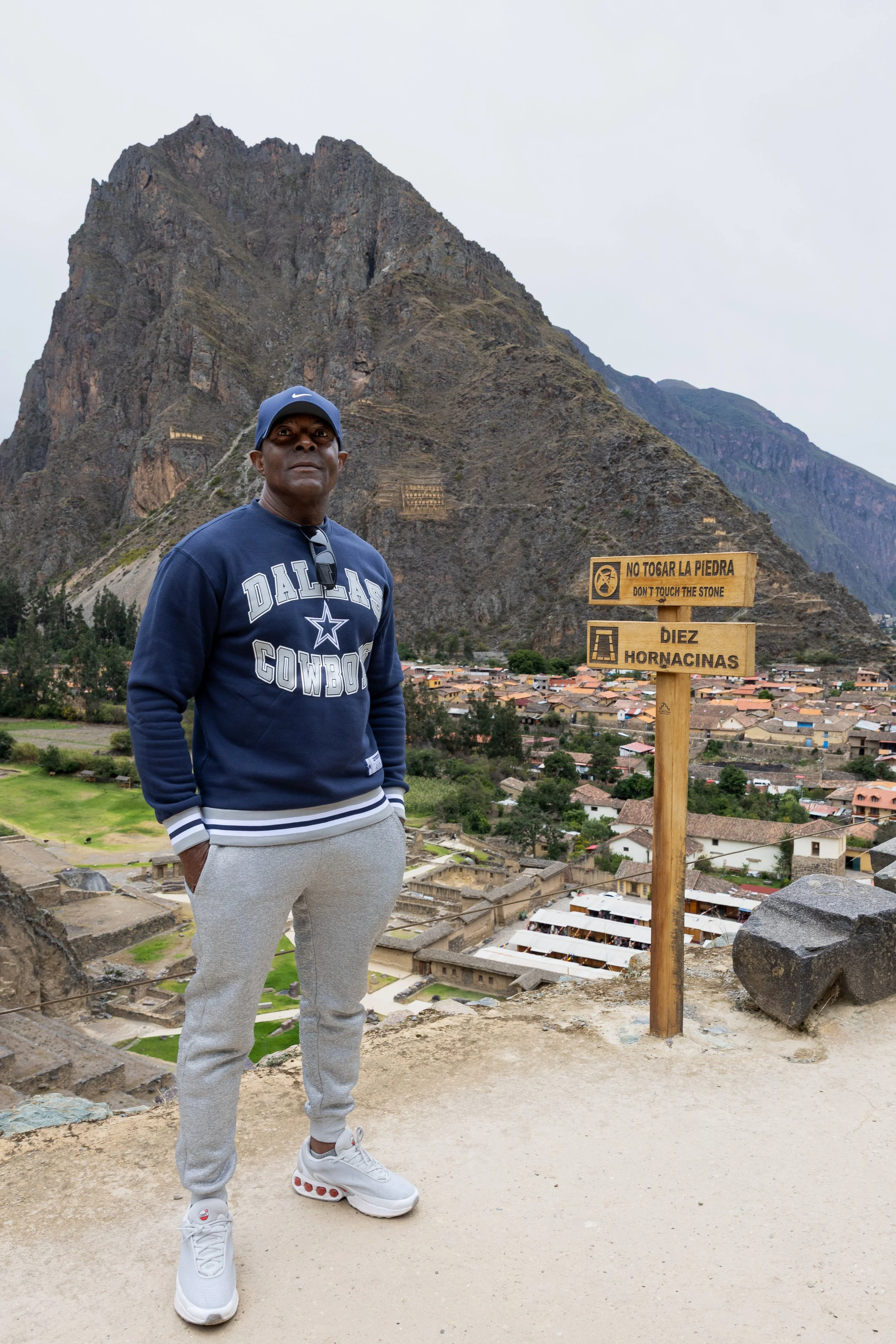 A man wearing a Dallas Cowboys sweatshirt, gray sweatpants, and white sneakers standing on a dirt path in front of a steep mountain with ancient ruins below. A sign next to him reads: "No Tocar La Piedra" and "Don't Touch the Stone" with another note