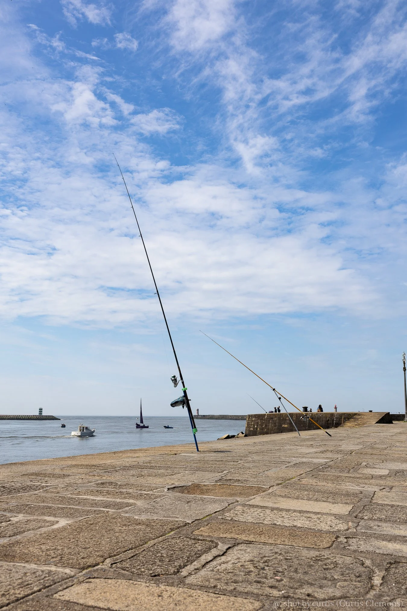 A harbor with three fishing rods set up on a paved walkway. There are boats and sailboats on the water, with a breakwater and people walking along the pier under a partly cloudy blue sky.