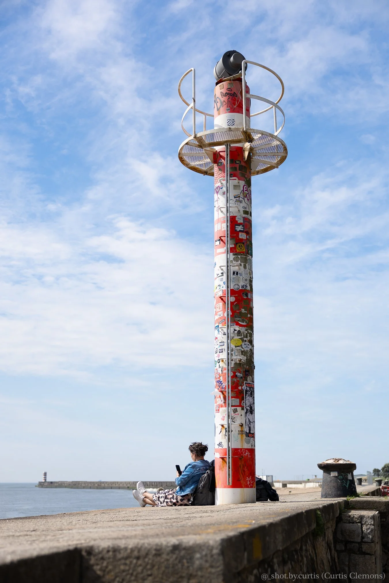 Person sitting on a concrete seaside promenade, wearing casual clothes and using a smartphone, next to a red and white graffiti-covered lighthouse with a lookout platform, under a blue sky with some clouds.