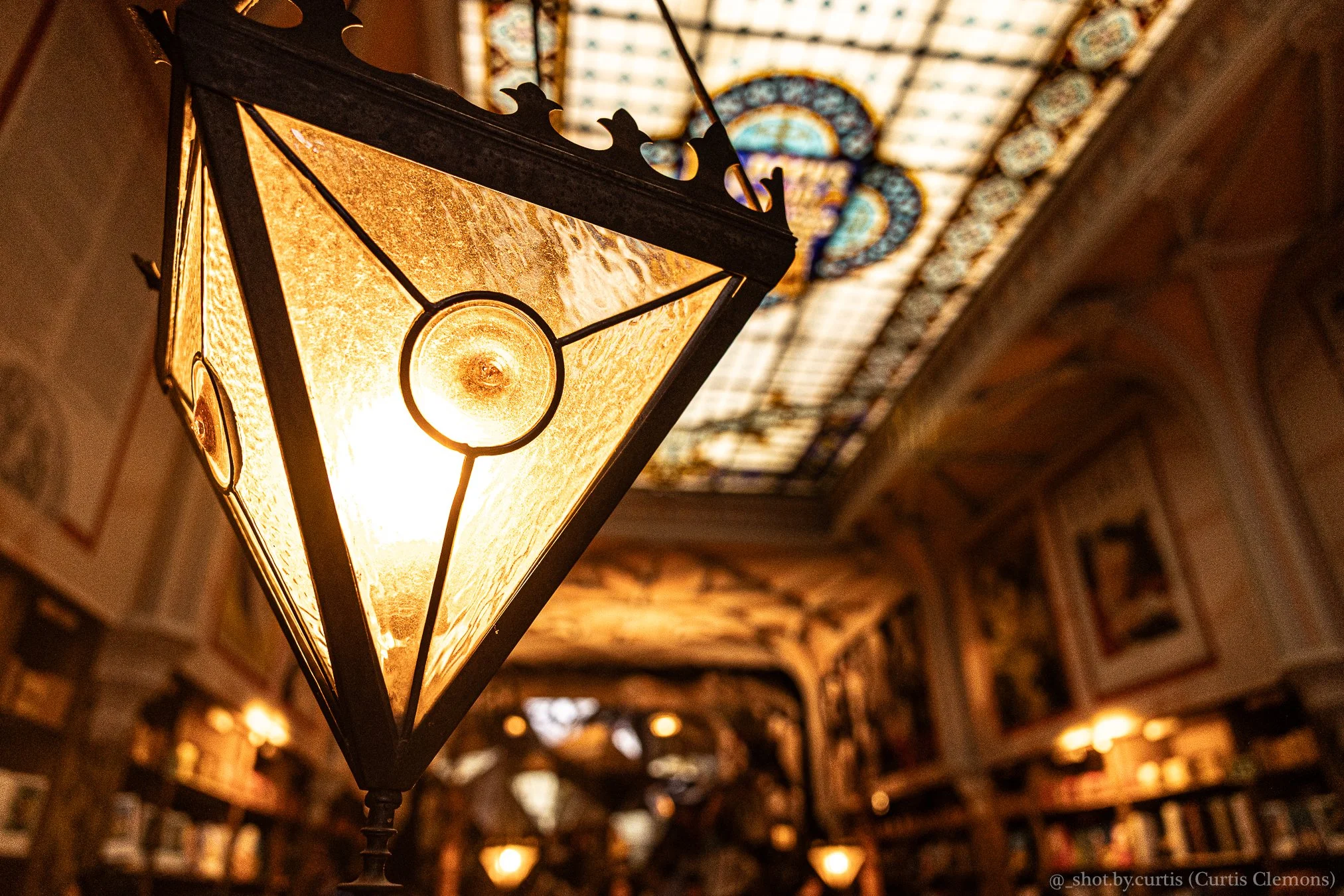 Close-up of an ornate, glowing lantern hanging from the ceiling inside a building with intricate architectural details and stained glass patterns in the background.