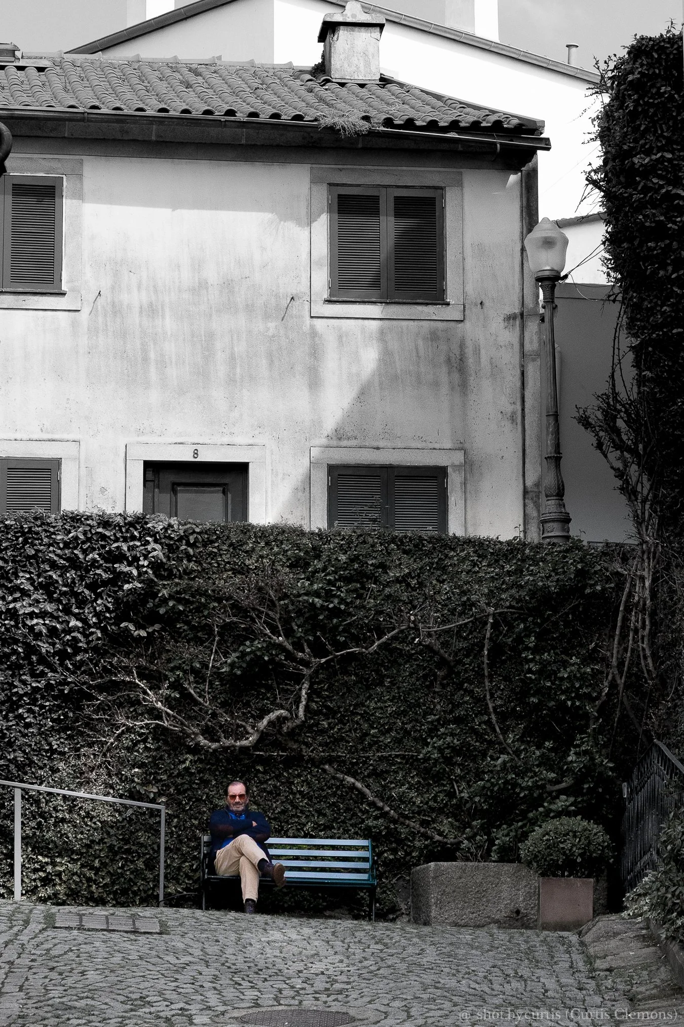 A man sitting on a bench in front of a bushy hedge, wearing sunglasses, a dark jacket, and beige pants. The background shows a building with multiple windows and a tiled roof, along with a lamp post on the right side.
