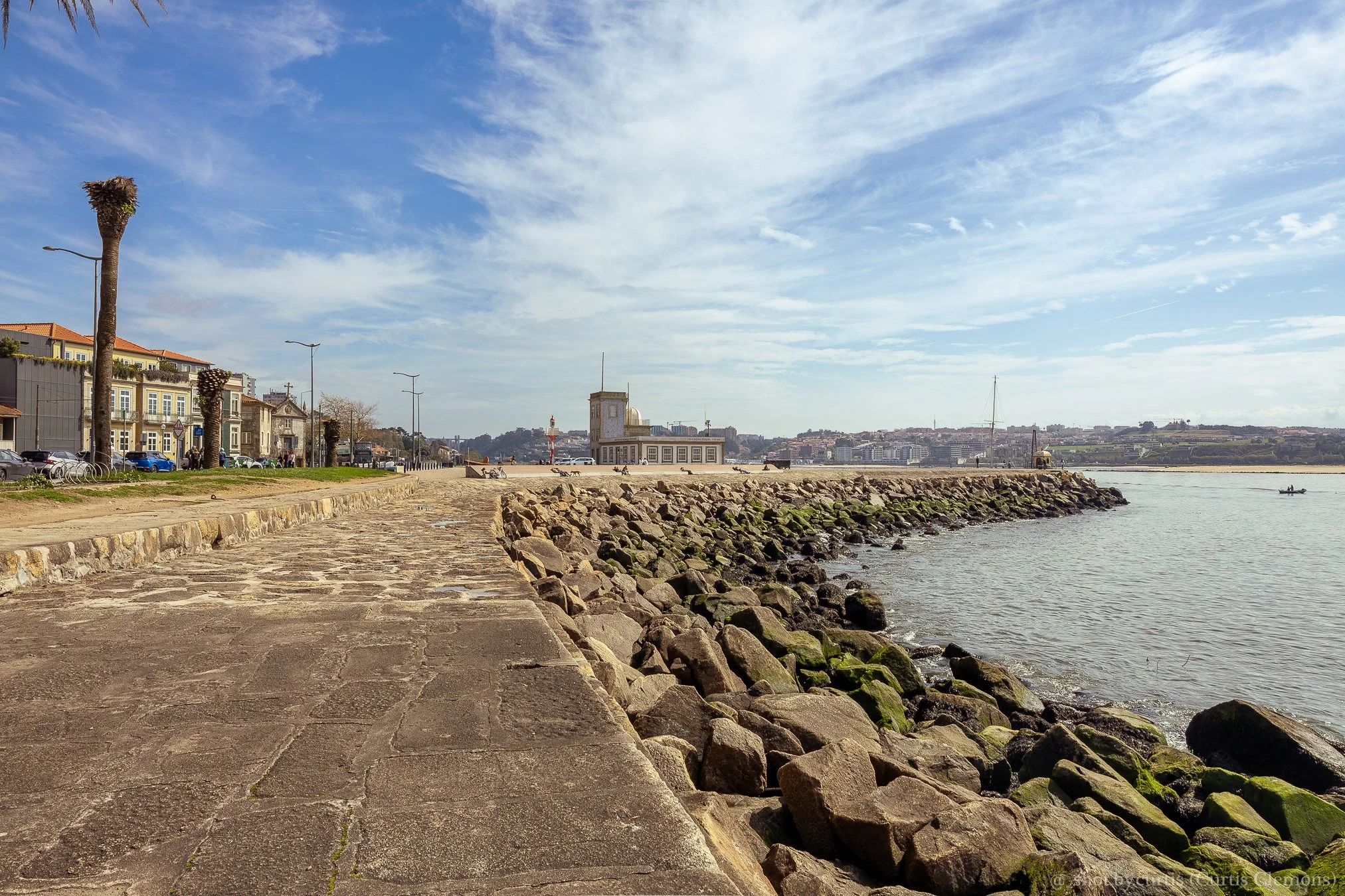 A waterfront scene with a stone walkway along the harbor, a rocky shoreline, boats on the water, and a building with a clock tower in the background under a partly cloudy sky.
