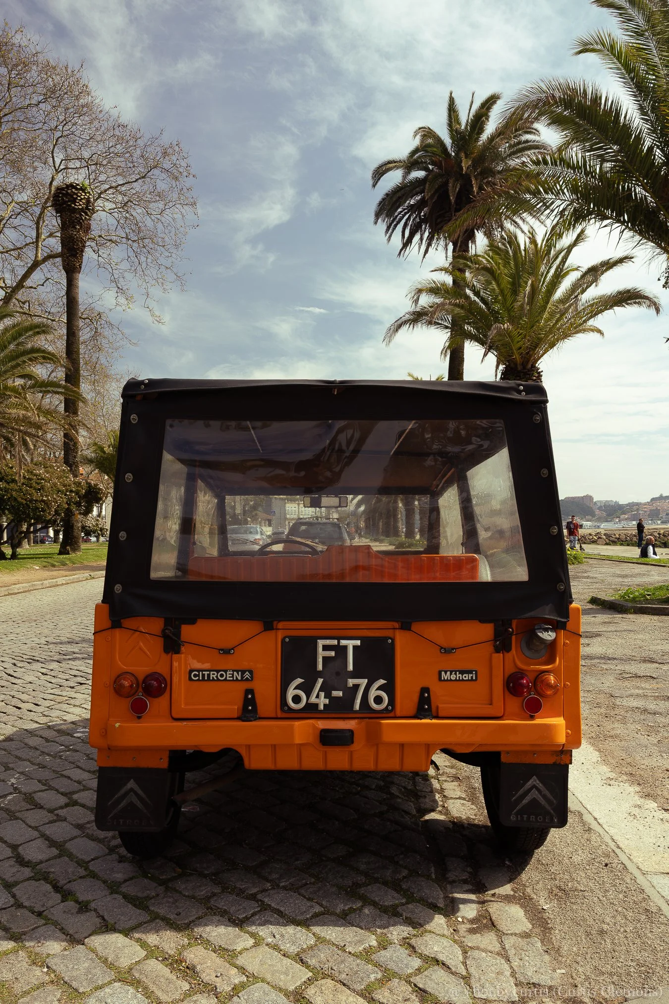Rear view of an orange Citroën Méhari vehicle with black soft top, showing classic tail lights, license plate 'FT 64-76,' and black mudguards, parked on a cobblestone street with palm trees and a partly cloudy sky in the background.