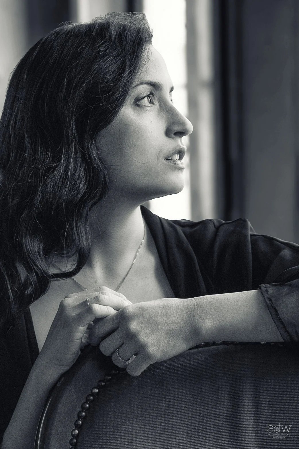 A black-and-white photo of a woman with long, wavy hair looking out a window, her hands resting on a piece of furniture, wearing a dark top and a necklace.