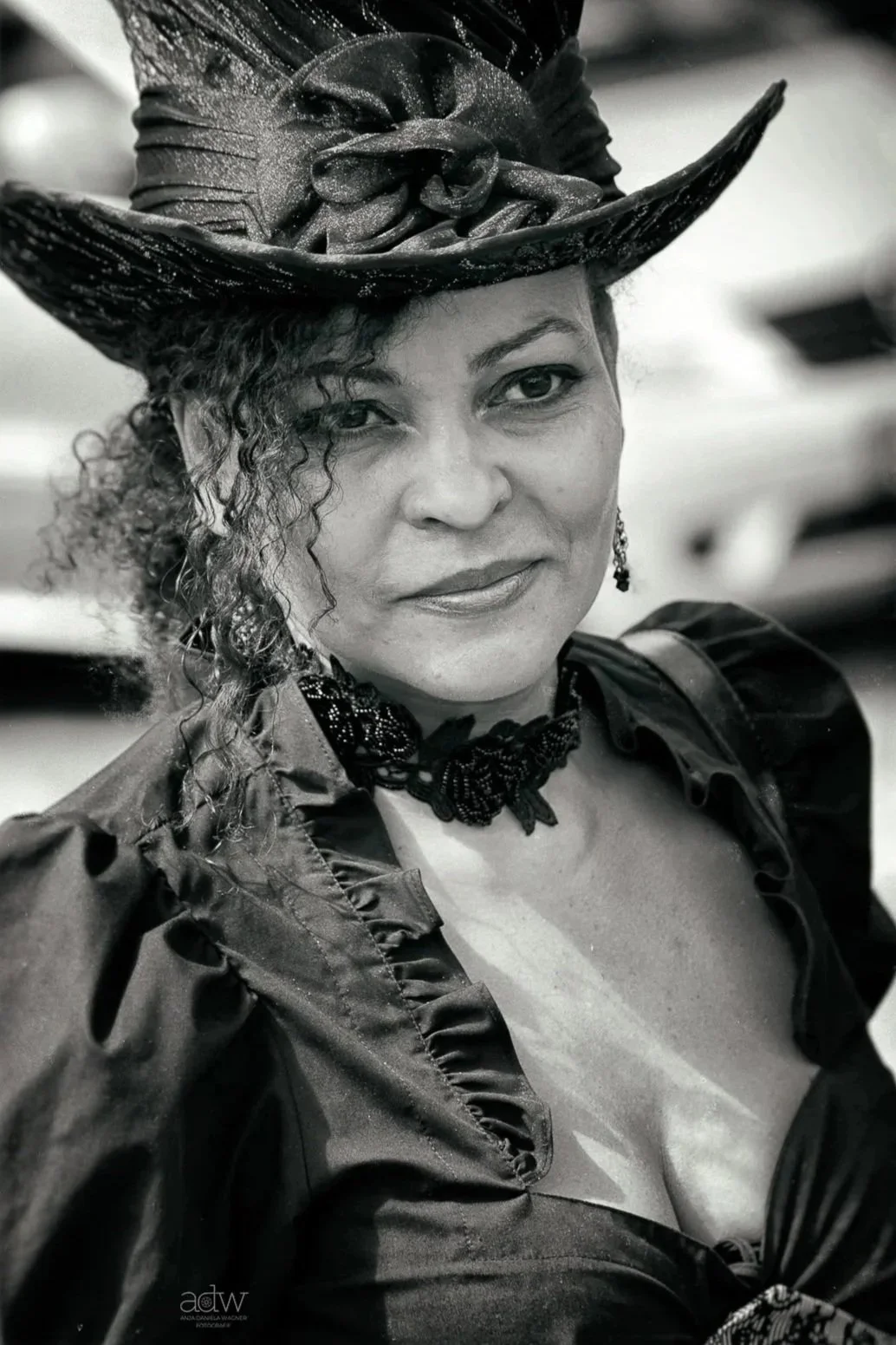 A woman wearing a fancy, large, decorated hat, and dark, ruffled clothing, smiling slightly, looking at the camera in black and white.