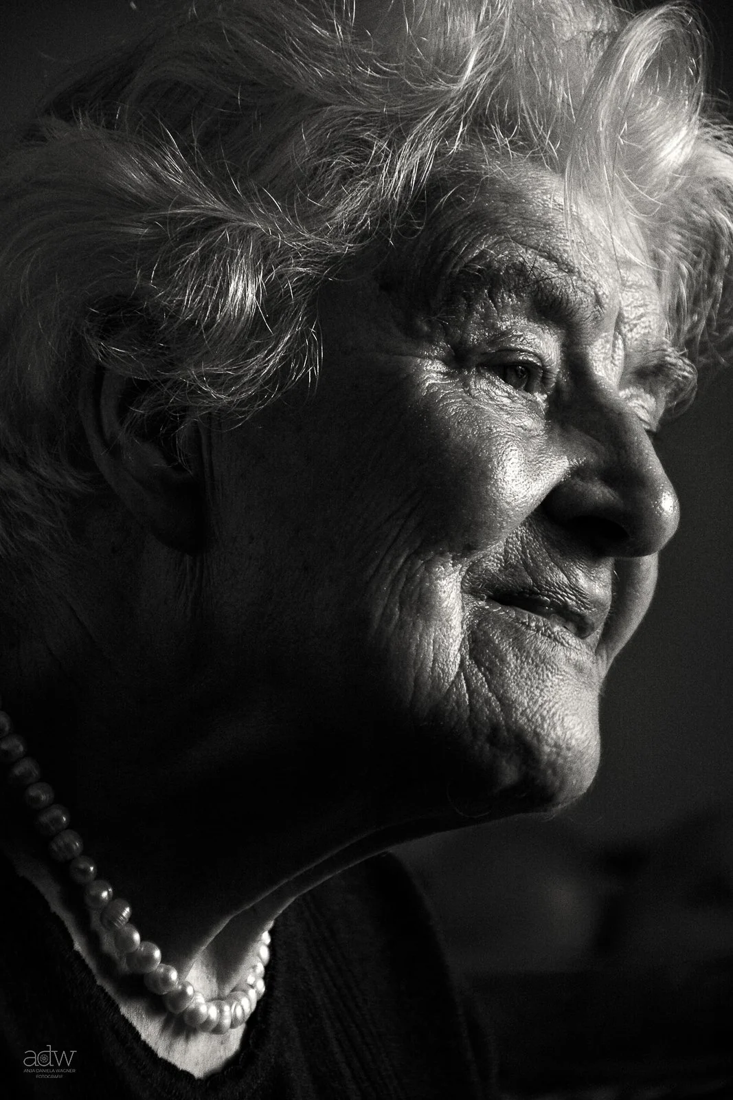 Close-up black and white portrait of an elderly woman with wrinkled skin and white hair, wearing a pearl necklace.