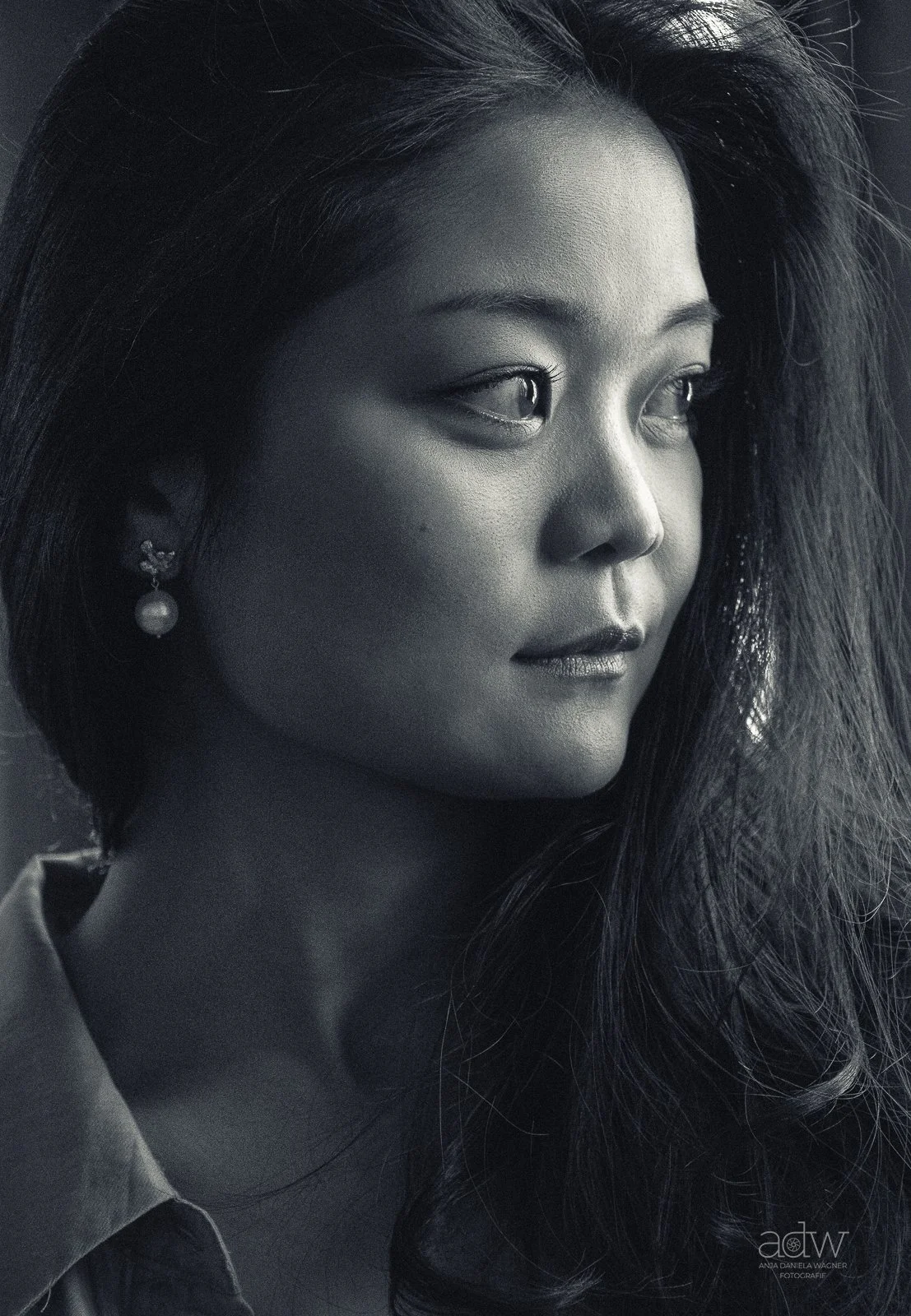 A black and white close-up portrait of a woman with long hair, earrings, and a serious expression, looking to the side.