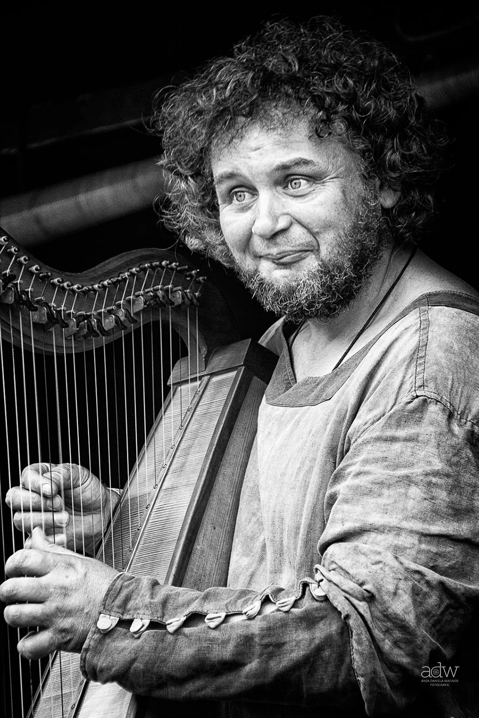 A black and white photo of a person with curly hair and a beard playing a harp, looking to the side with a gentle expression, wearing a long-sleeved shirt.