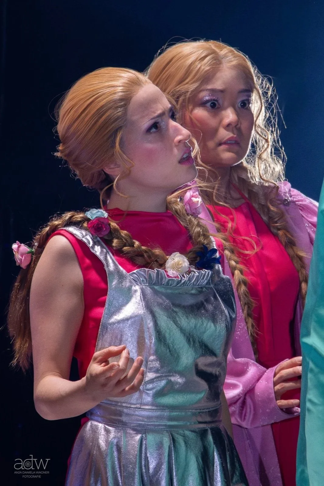 Two women with long braided blond hair dressed in vivid pink and shiny outfits look with concern at something off-camera.