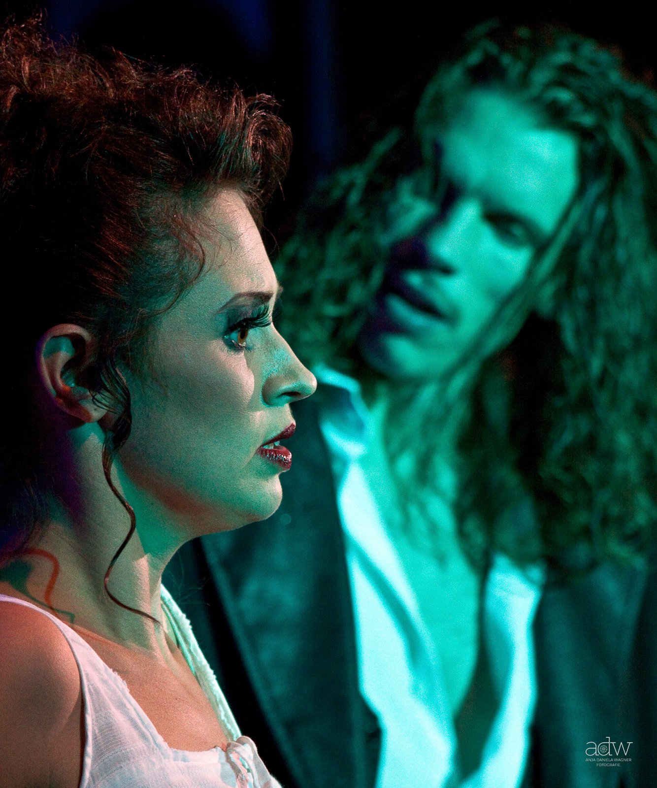 A woman with styled hair and makeup looking at a screen displaying a man with long curly hair, both illuminated by teal lighting.