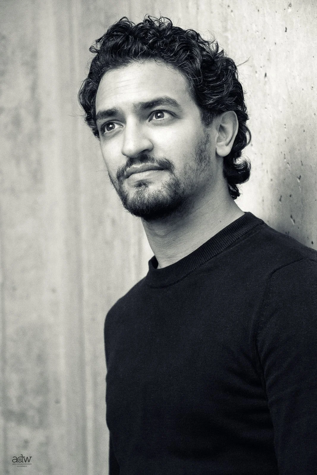 Black and white portrait of a man with curly hair, a beard, and a mustache, wearing a dark shirt, standing against a plain wall, looking thoughtfully to the side.