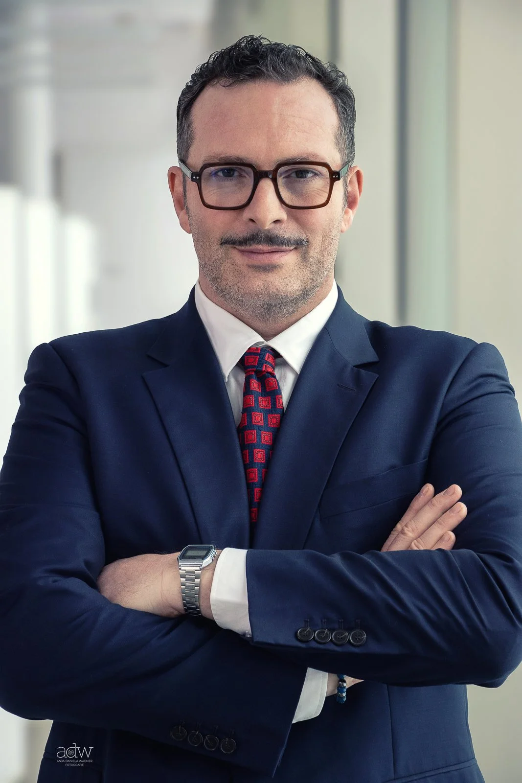 A confident man in a navy suit, white shirt, red patterned tie, and glasses, standing with arms crossed in a modern office setting.