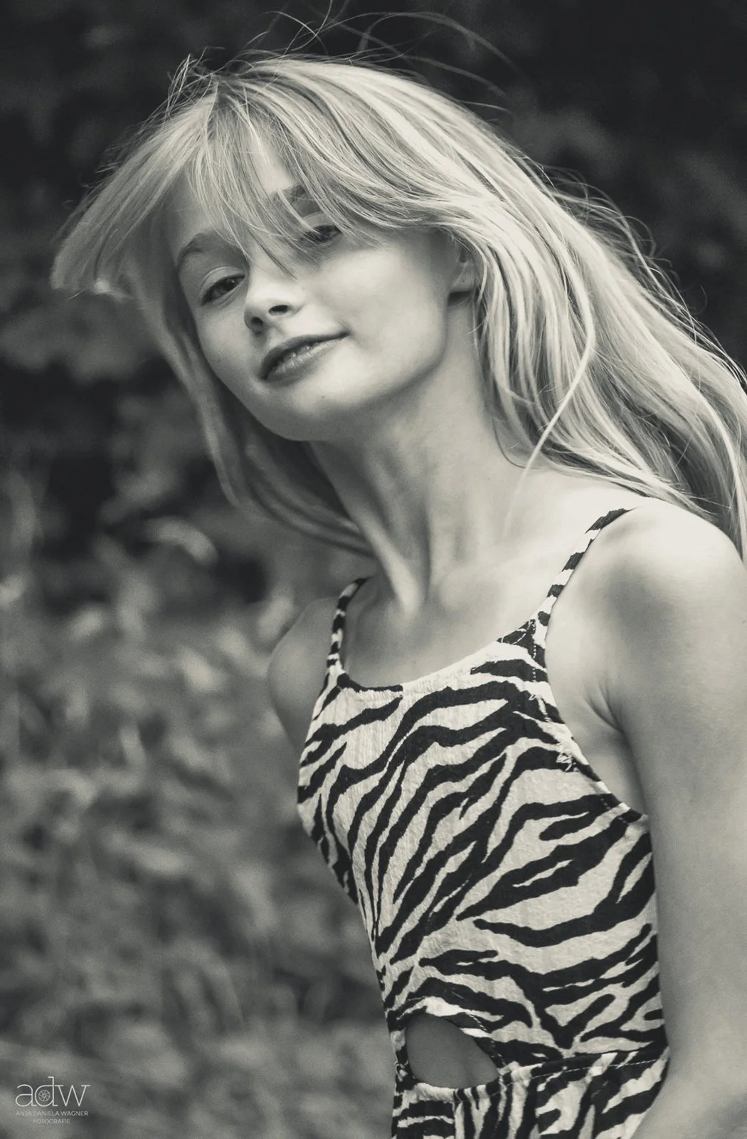 Black and white photo of a young woman with long, tousled blonde hair, smiling softly, wearing a sleeveless zebra print top, outdoors with blurred foliage in the background.