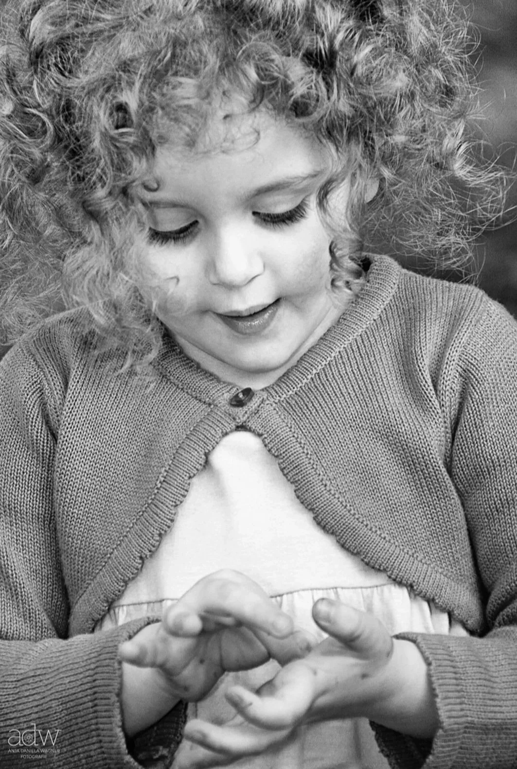 A young girl with curly hair looking down and smiling, wearing a cardigan and a white top, with her hands clasped together.
