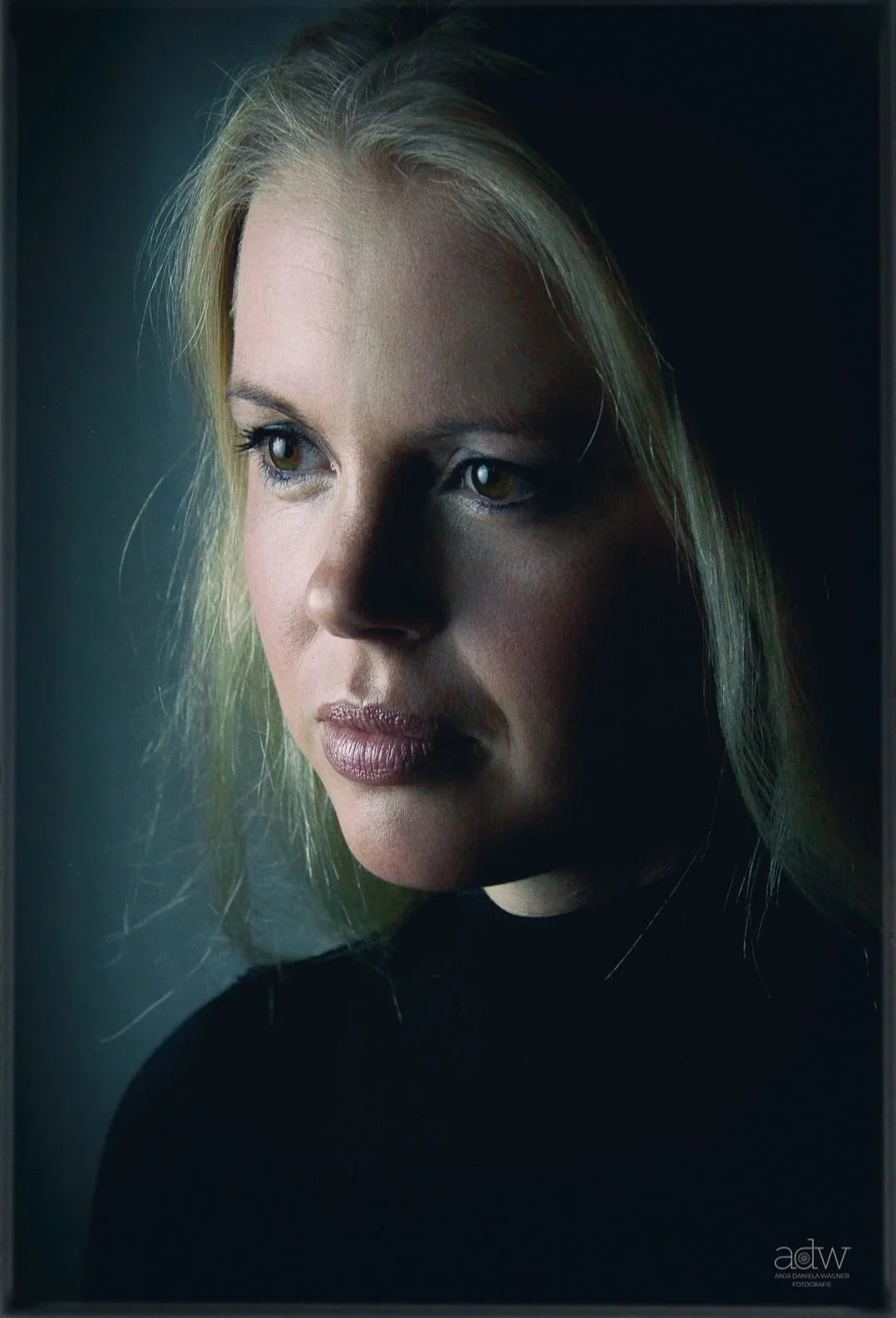 Close-up portrait of a woman with blonde hair wearing a black top, gazing to the left with a thoughtful expression, against a dark background.