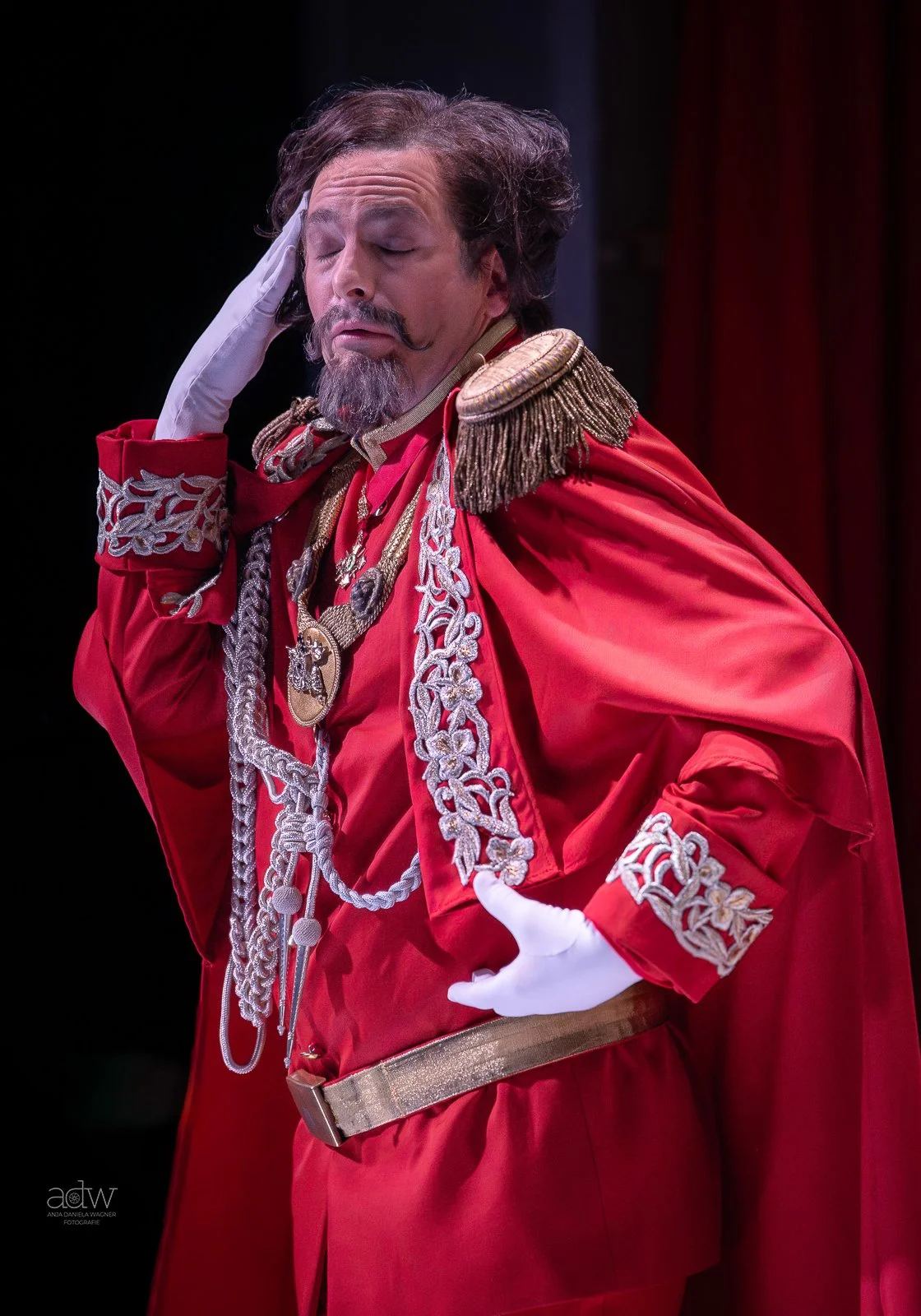 A man in a red military-style costume with gold and silver embellishments, wearing a white glove, with a hand on his forehead, eyes closed, and a thoughtful expression.