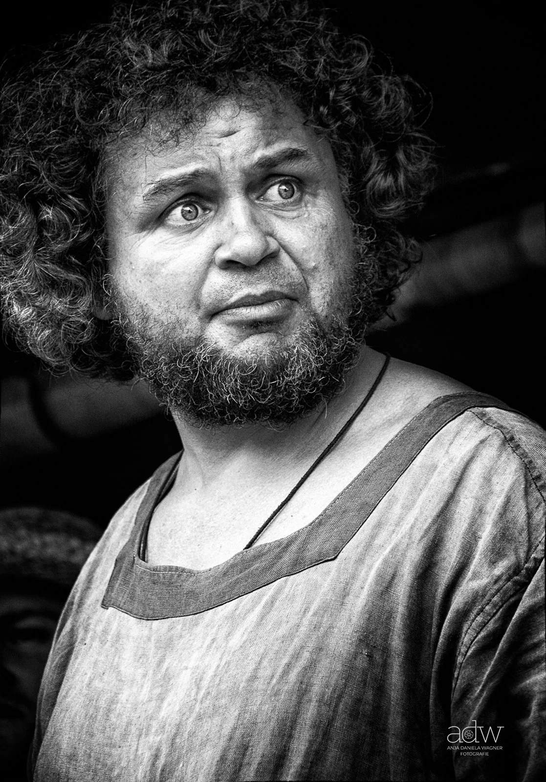 Black and white portrait of a man with curly hair and a beard, wearing a traditional shirt and necklace, looking upwards.