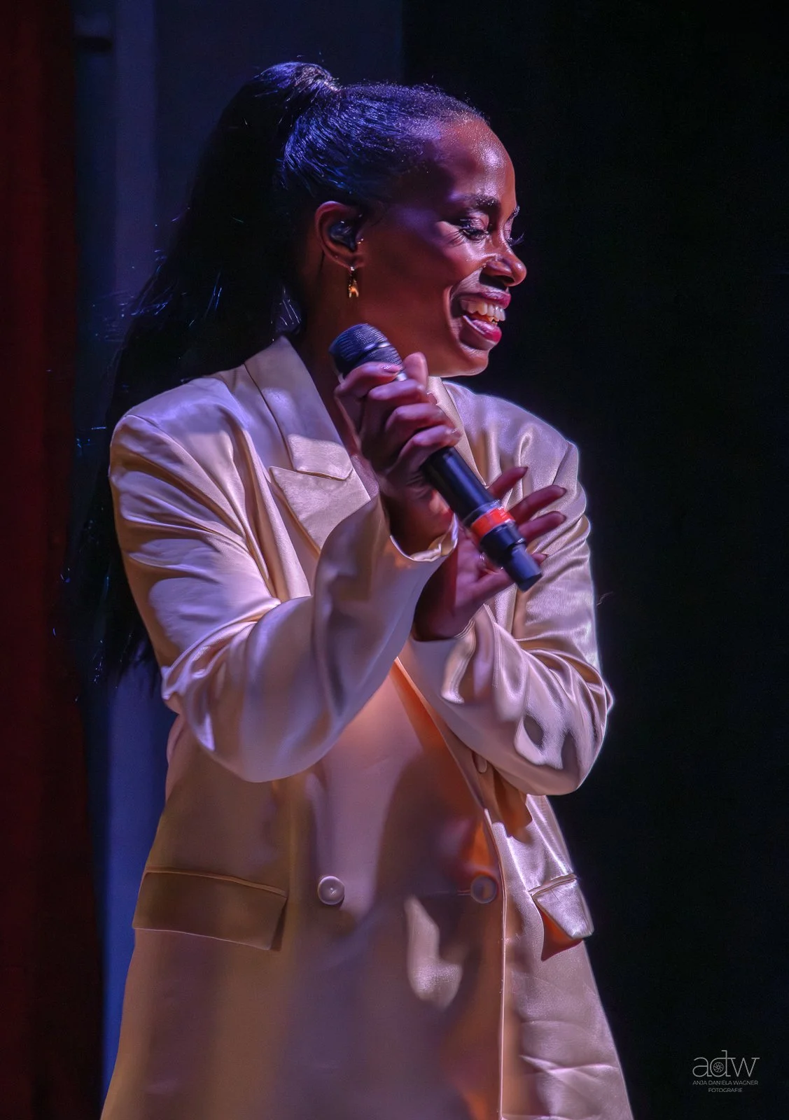 A woman smiling and holding a microphone on stage, dressed in a shiny beige blazer.