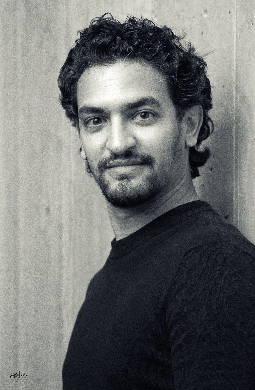 Black and white portrait of a man with curly hair, a beard, and mustache, wearing a dark shirt, standing against a wooden wall, looking at the camera with a slight smile.