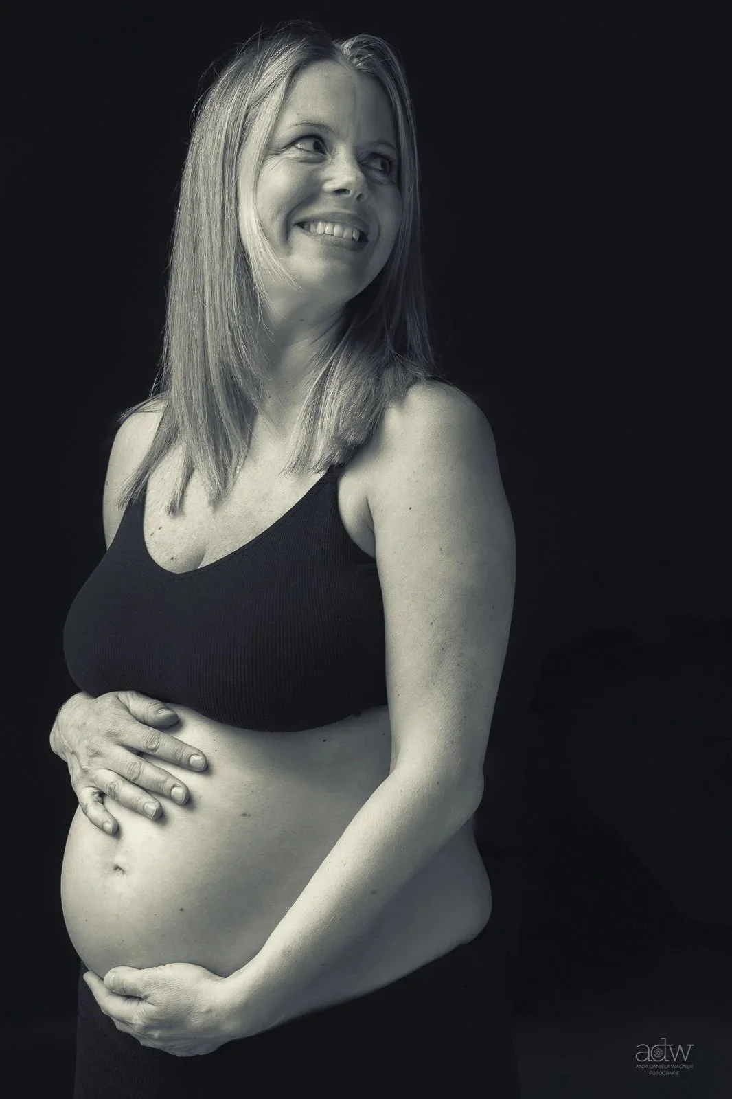 Black and white portrait of a pregnant woman smiling and looking to her right, with her hand resting on her belly, wearing a black sleeveless top, against a dark background.