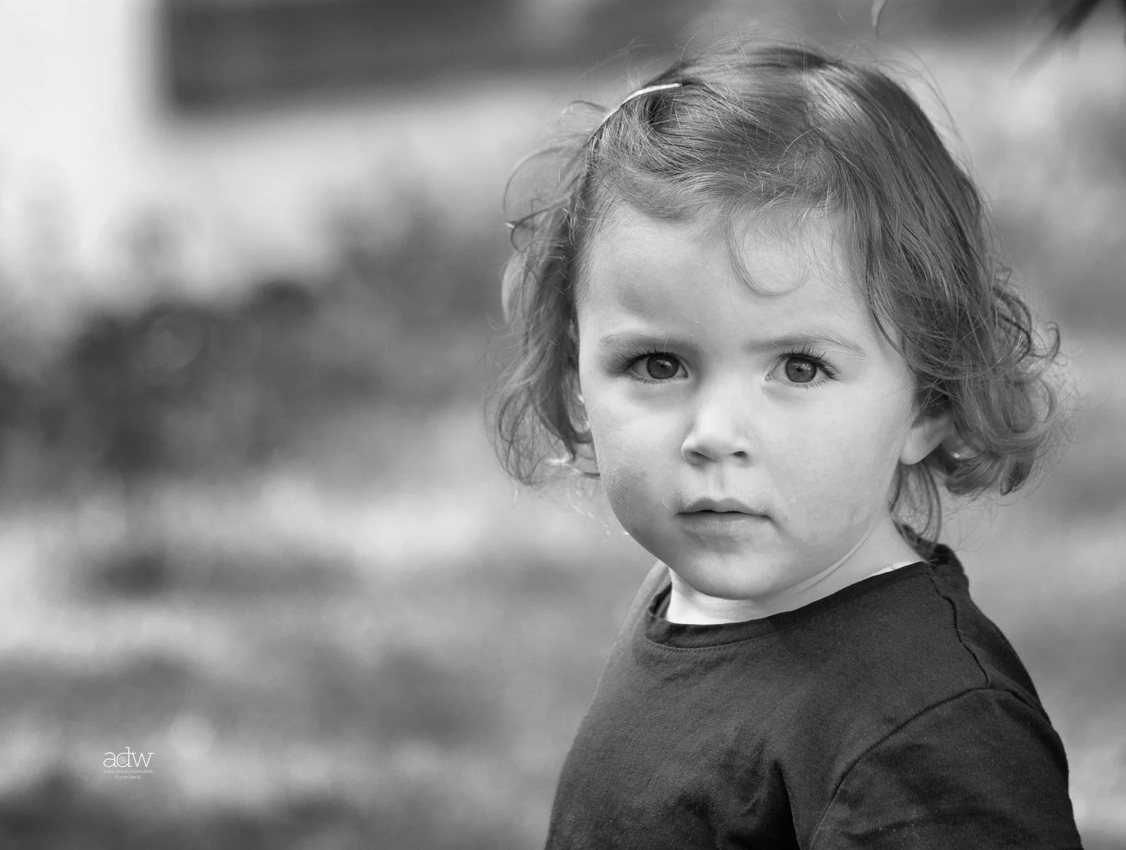 A young child with curly hair and light skin looking directly at the camera, wearing a dark shirt, outdoors with a blurred background.