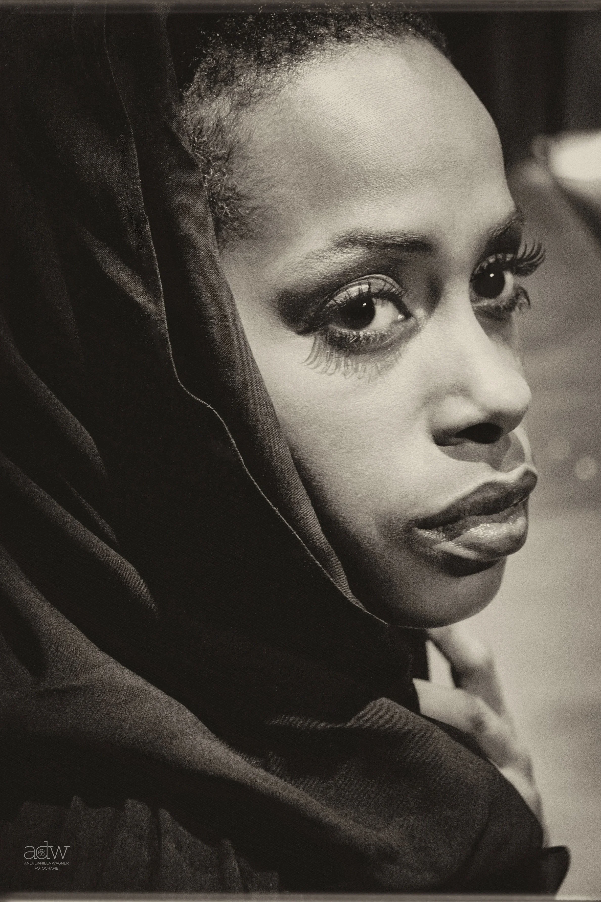 Close-up black and white portrait of a woman with smooth skin, large expressive eyes with highlighted eyelashes, full lips, and short natural hair, wrapped in dark fabric covering her head.