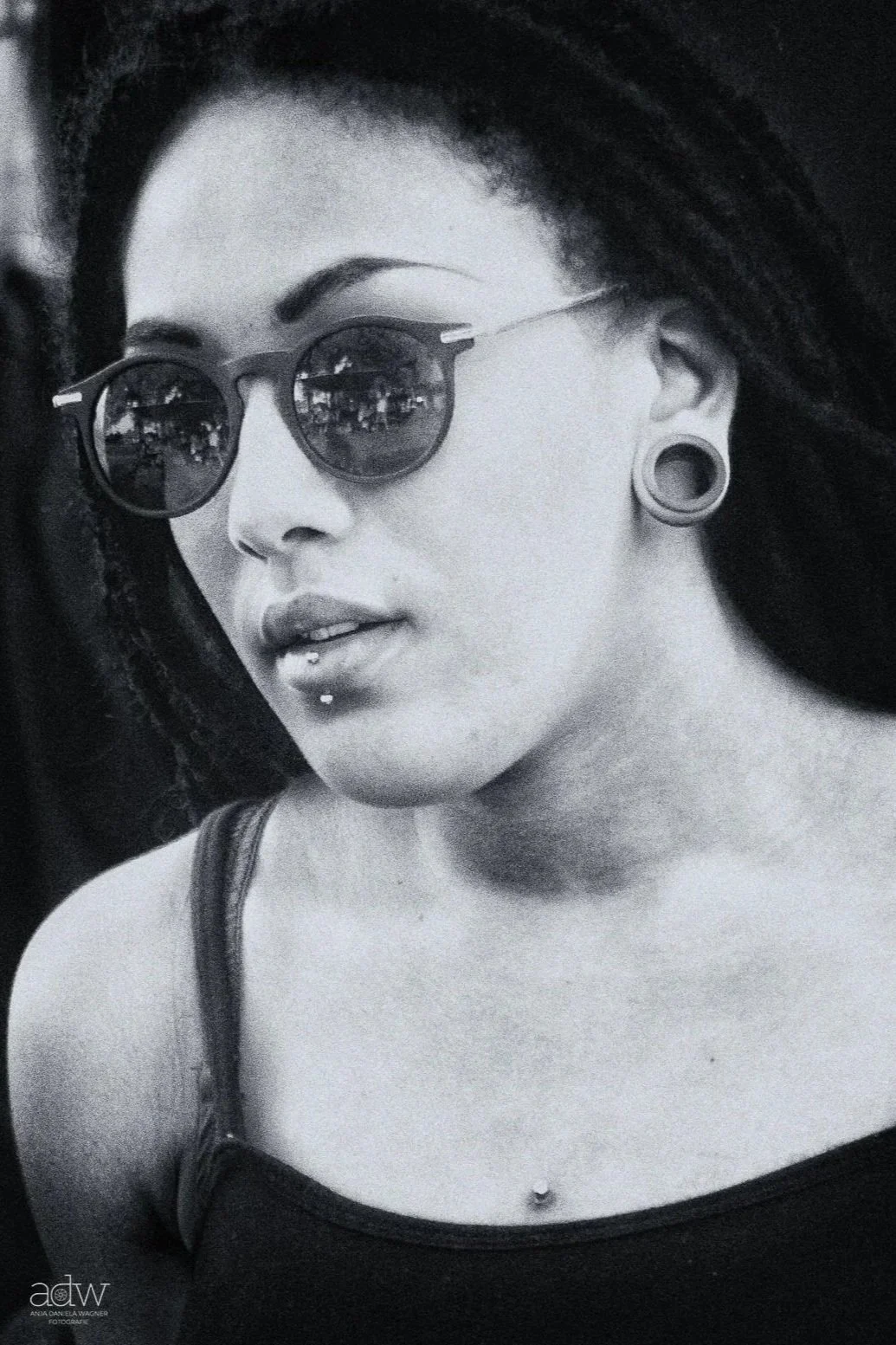 A black and white photo of a woman with tattoos, piercings, and large gauge earrings, wearing sunglasses, with long dark hair, and a tank top.