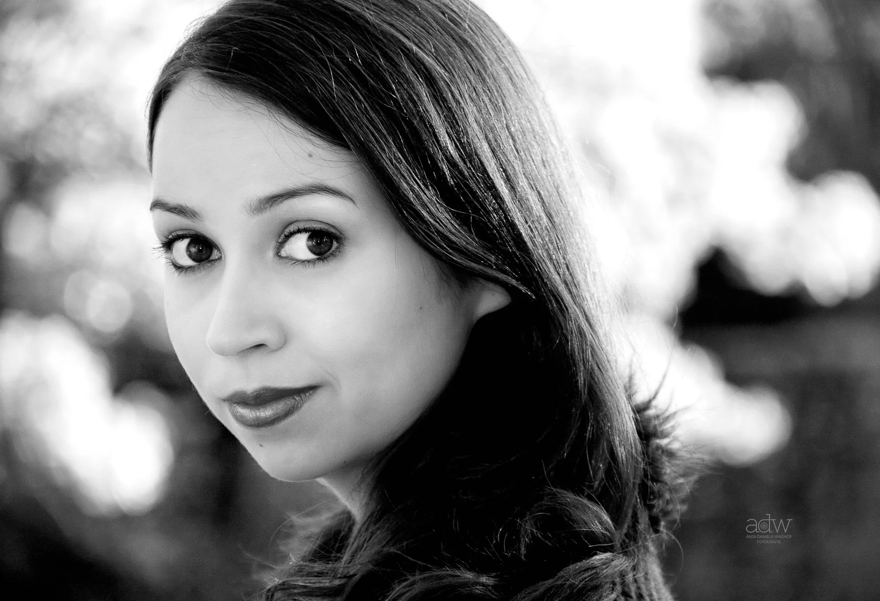 A close-up black and white portrait of a woman with dark hair, lightly smiling, with blurred background lights.