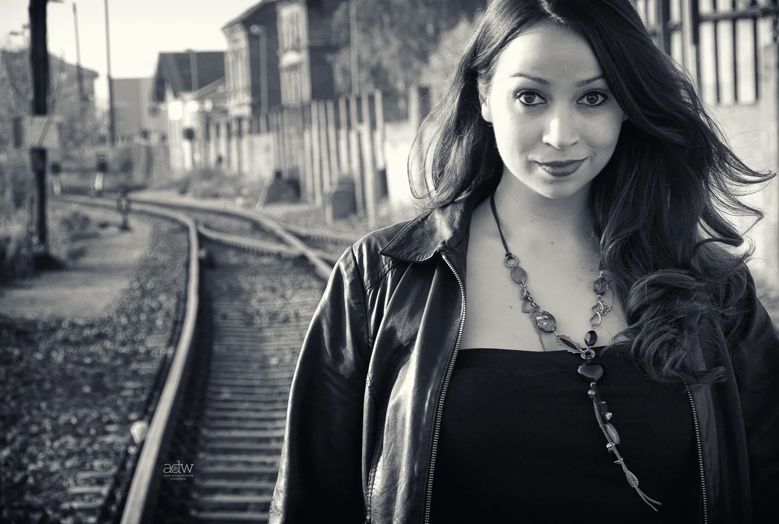 A woman with long dark hair wearing a leather jacket and necklace standing outdoors near train tracks and old buildings.