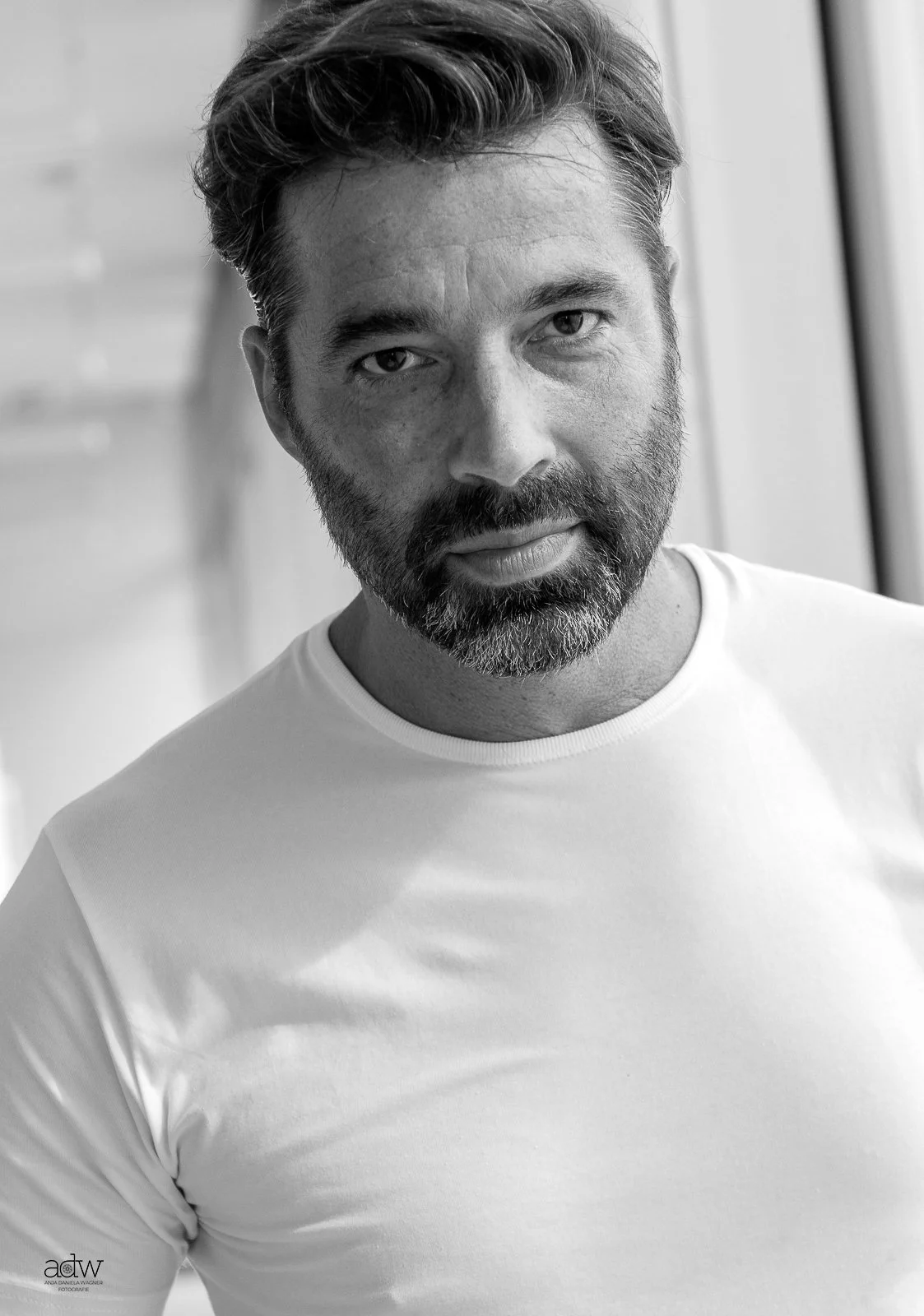 Black and white portrait of a man with a beard and short hair, wearing a white t-shirt, looking directly at the camera.