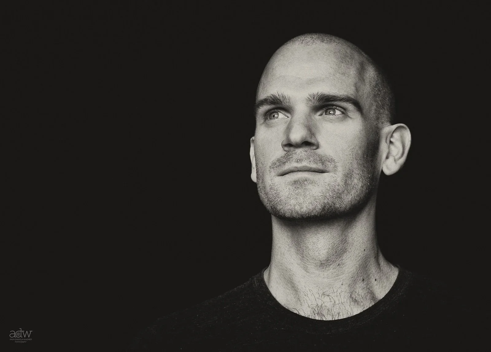 Black and white portrait of a man with a shaved head and short beard, wearing a dark shirt, against a dark background, looking slightly upward.
